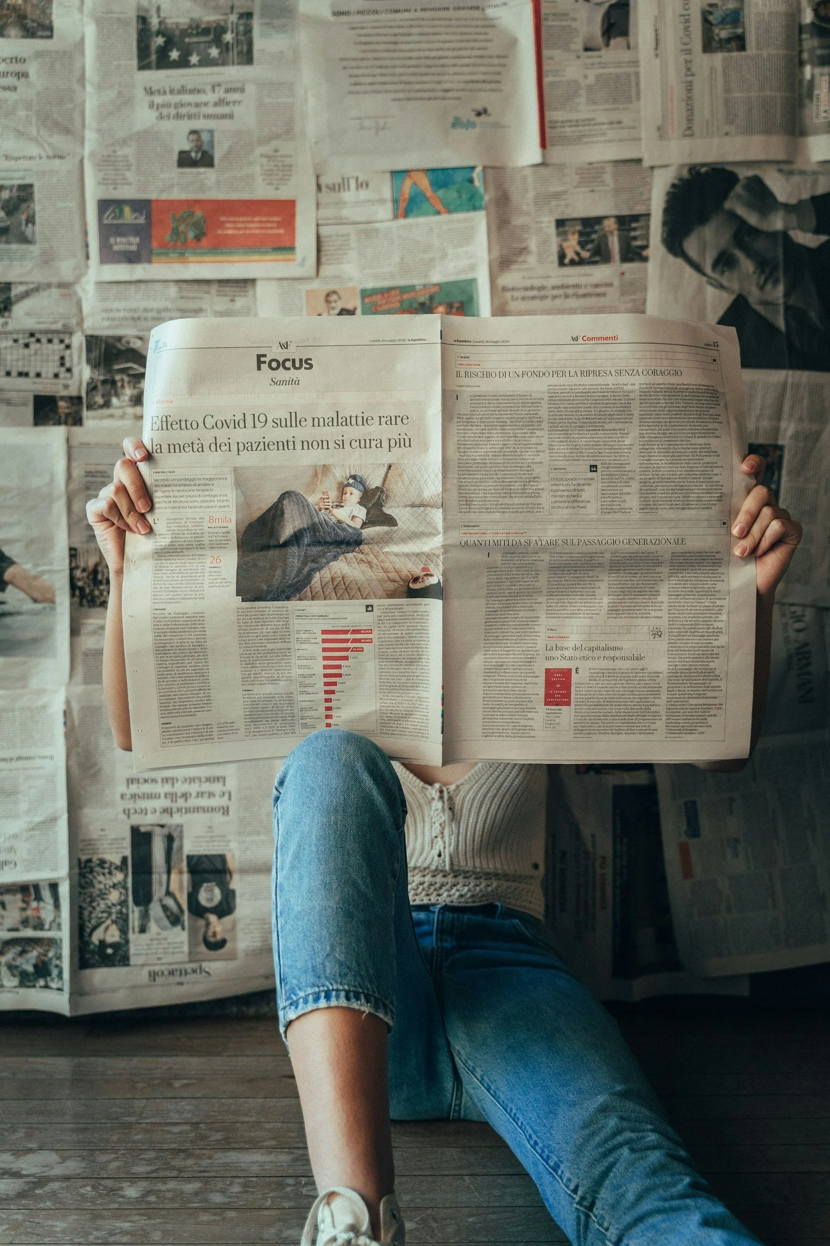 A person sitting on a wooden floor reading a newspaper, surrounded by scattered newspapers. The person is dressed in blue jeans and a striped shirt, with one knee bent and the other leg extended. The focus is on the newspaper content, which includes headlines and articles about COVID-19 and other topics.