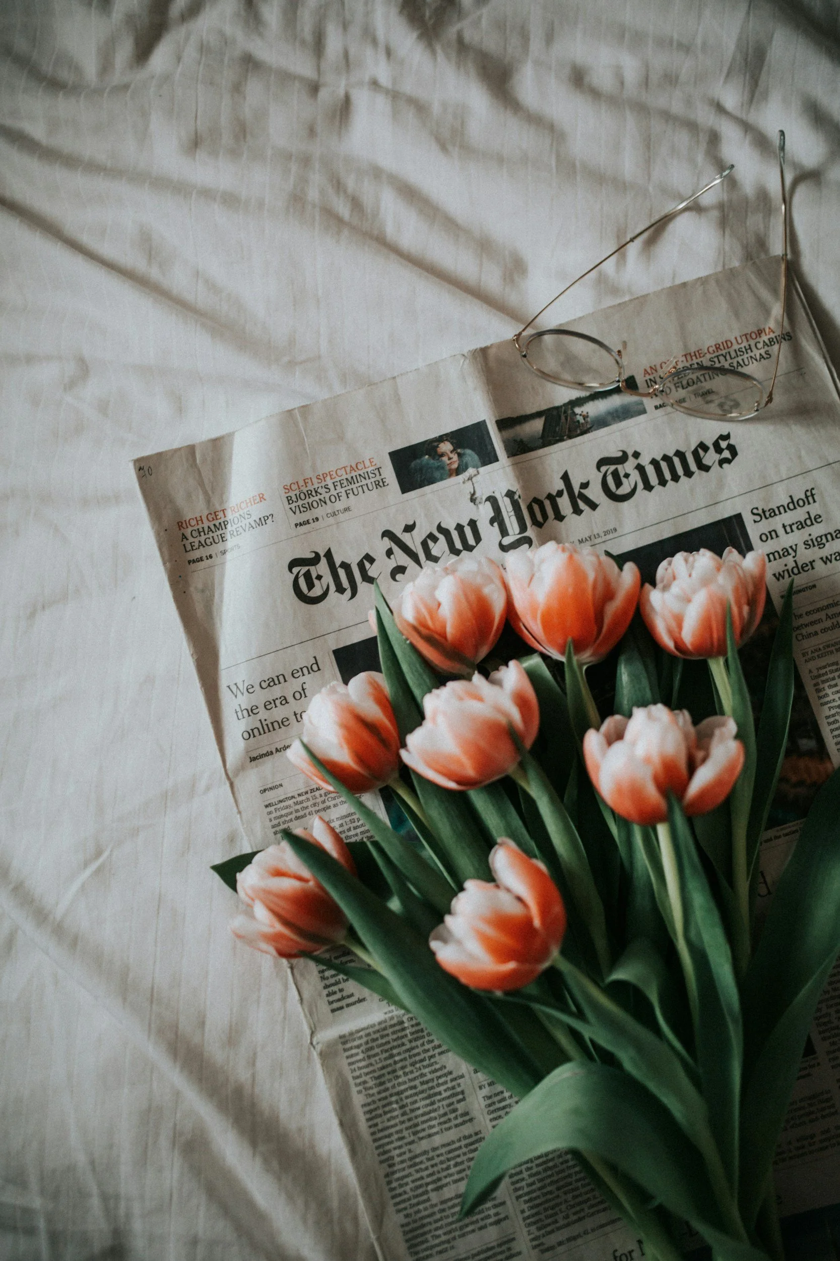 A bunch of pink and white tulips on top of a folded newspaper, with a pair of reading glasses resting on the newspaper.