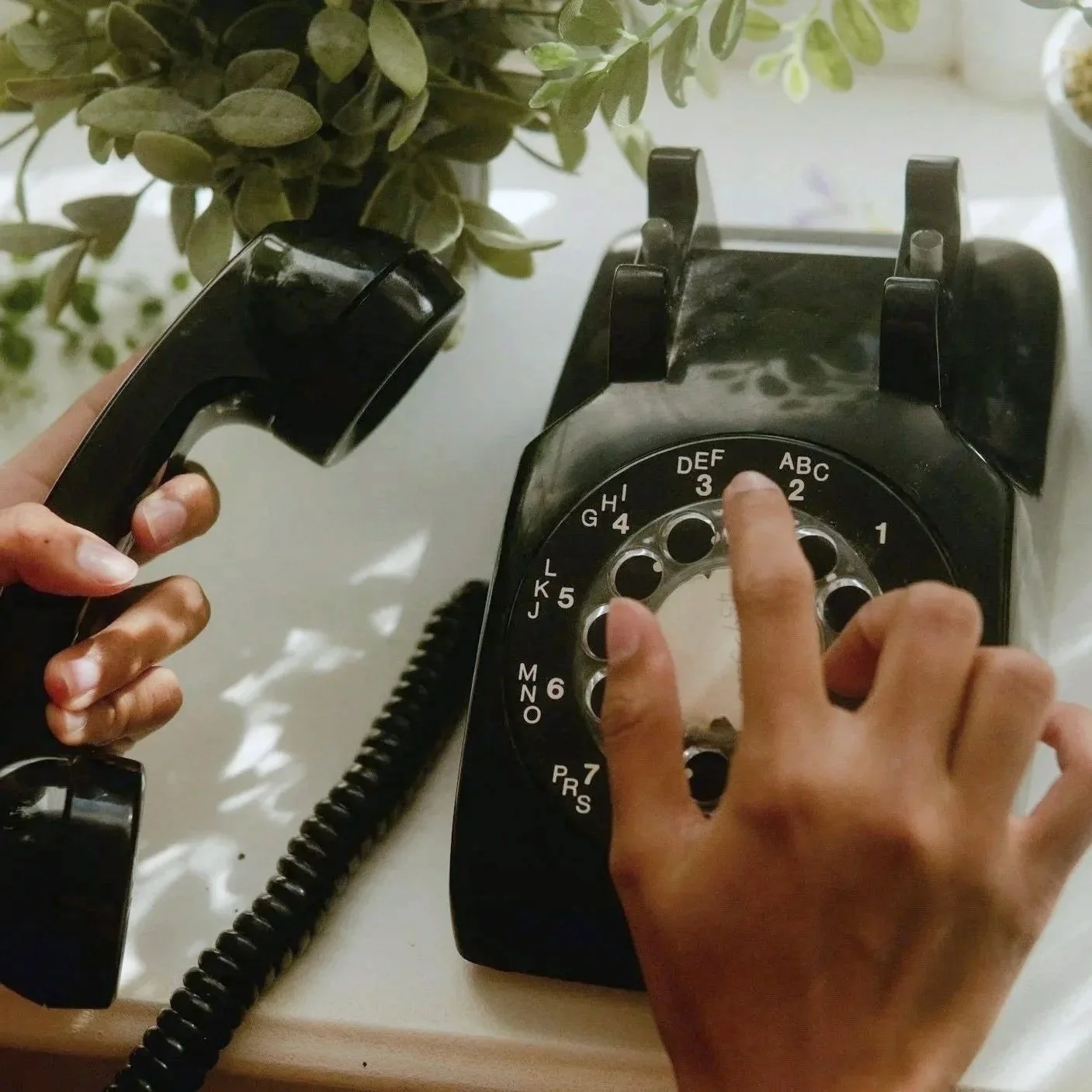 A person dialing a rotary phone with the finger on the number two, while holding the receiver in the other hand near a green potted plant.
