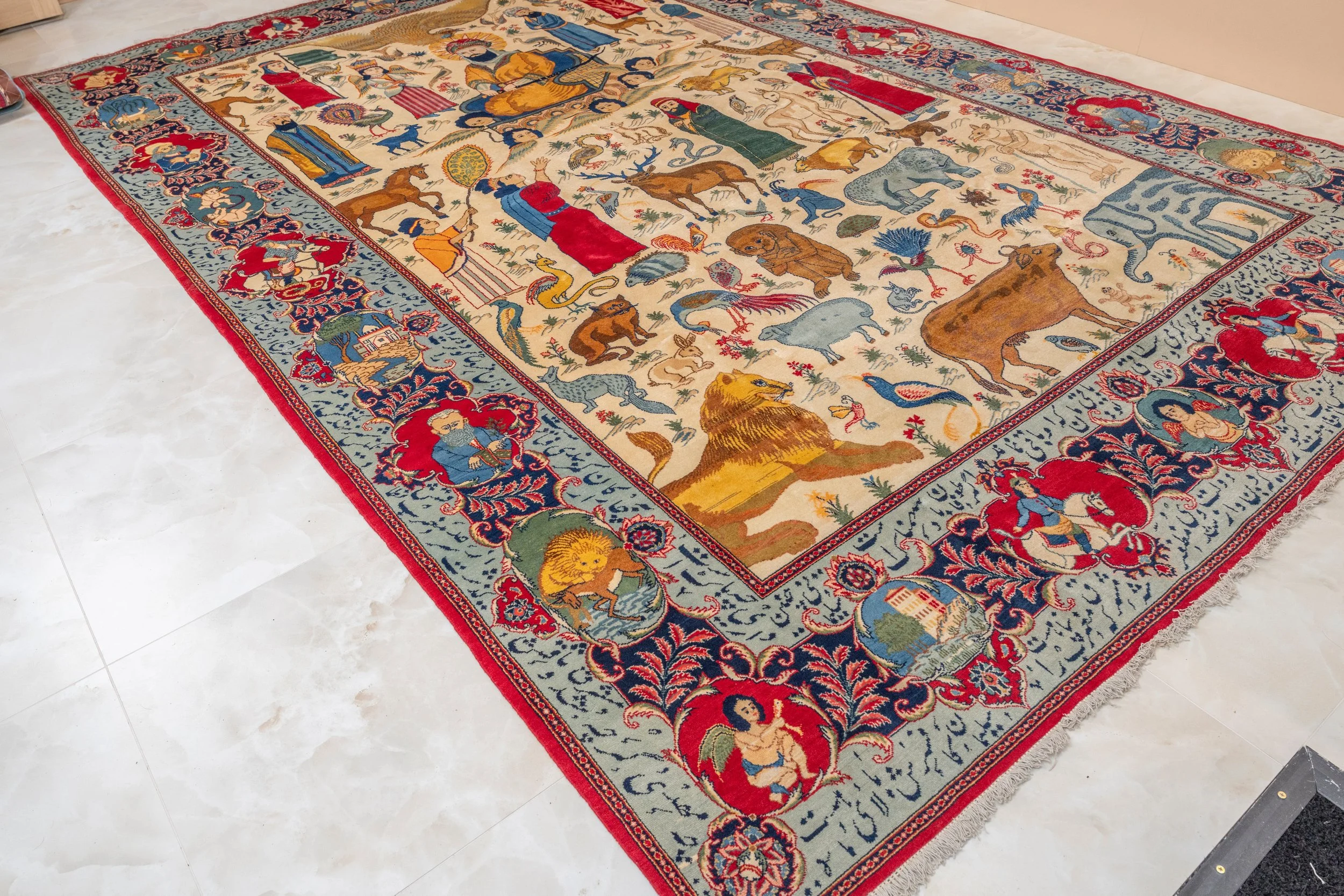 An intricately designed Tabriz Persian rug featuring animals, human figures, and floral patterns with a decorative border. The rug has a cream background with multicolored motifs including lions, elephants, deer, birds, and human portraits.