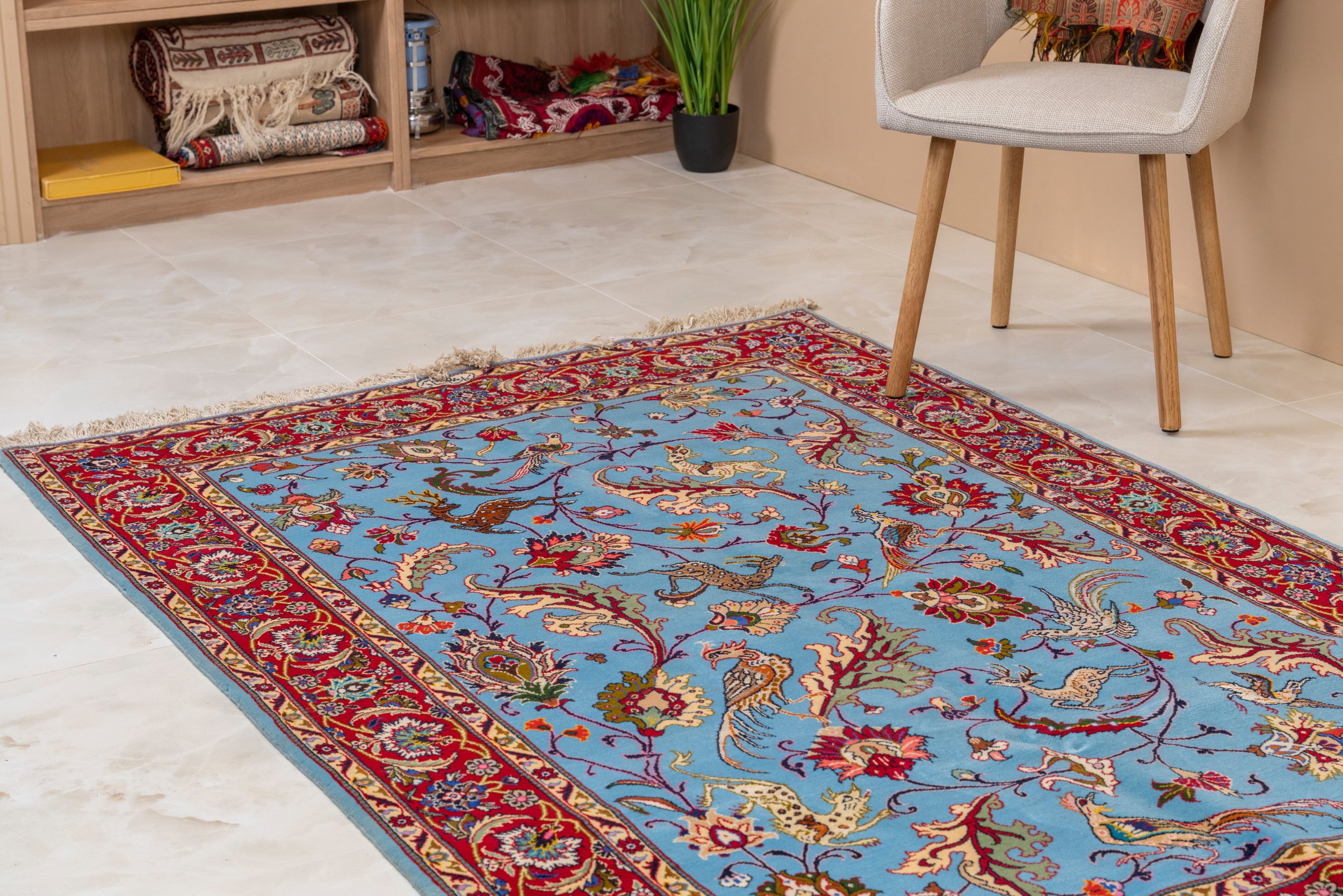 A patterned area rug with a blue background, red and green border, and intricate floral and bird designs, placed on a light-colored tiled floor in a room with shelves, a chair, and a potted plant.