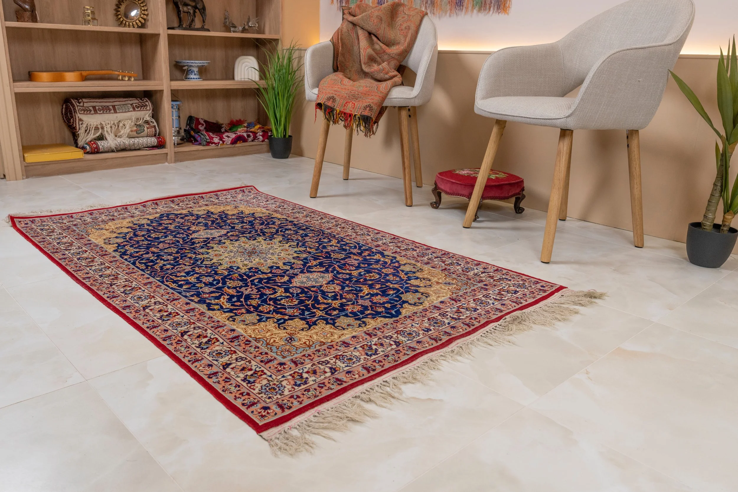 A colorful Persian-style rug with intricate blue, gold, and red patterns on a beige tiled floor in a cozy room with white chairs, potted plants, and shelves with folded textiles and decorative items.