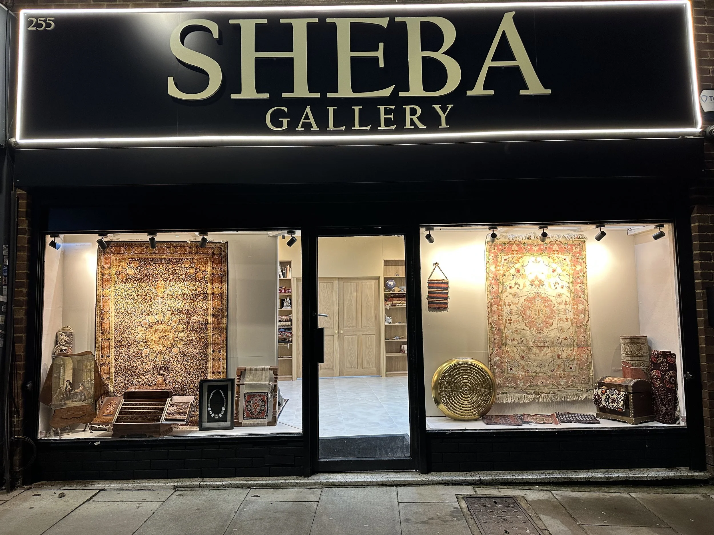 View of Sheba Gallery storefront at night with illuminated sign, displaying Persian rugs, jewelry, and decorative items in the window display.