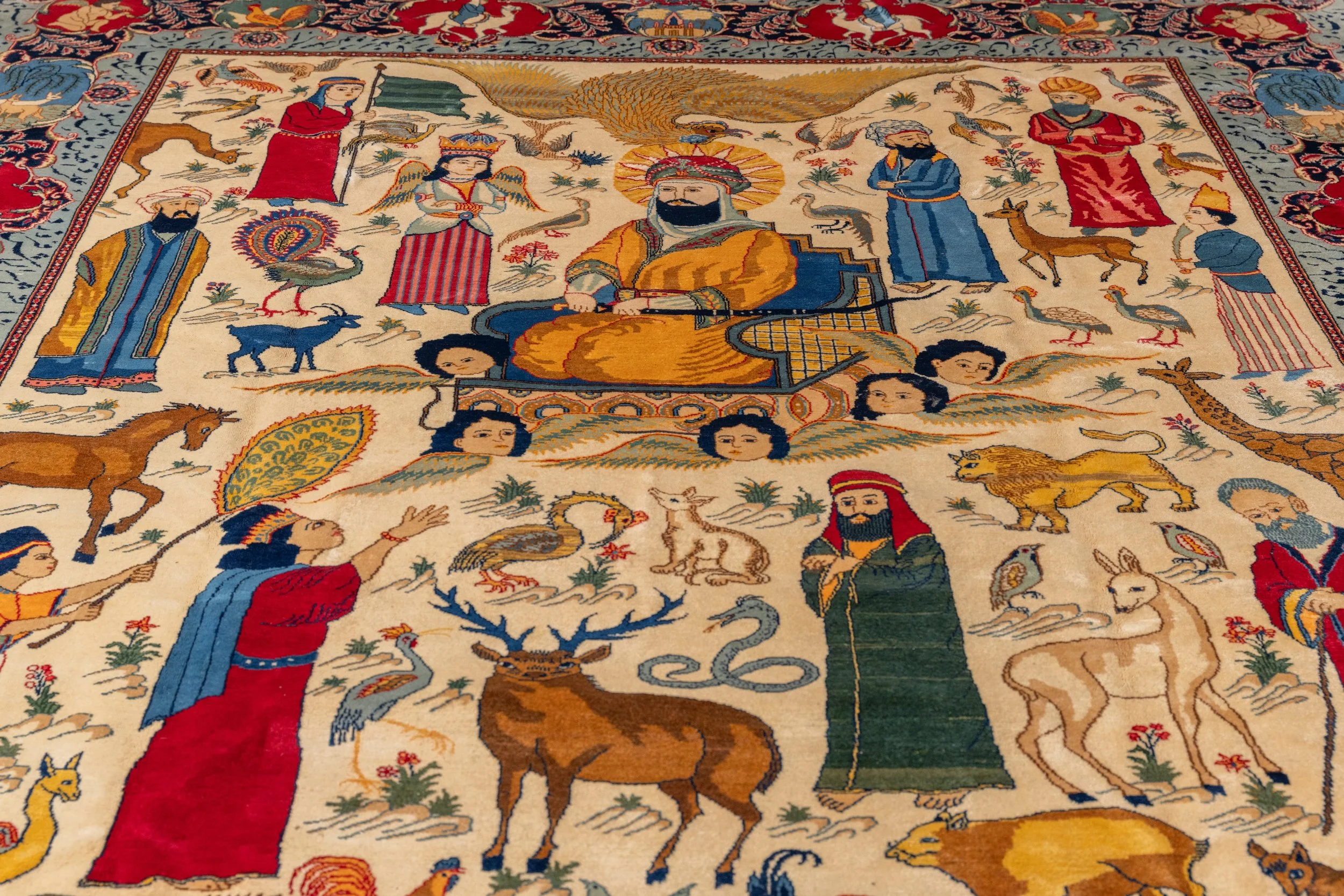 Colorful Persian-style rug depicting a central figure with a halo, surrounded by animals, birds, and multiple human figures, in a detailed and intricate design.