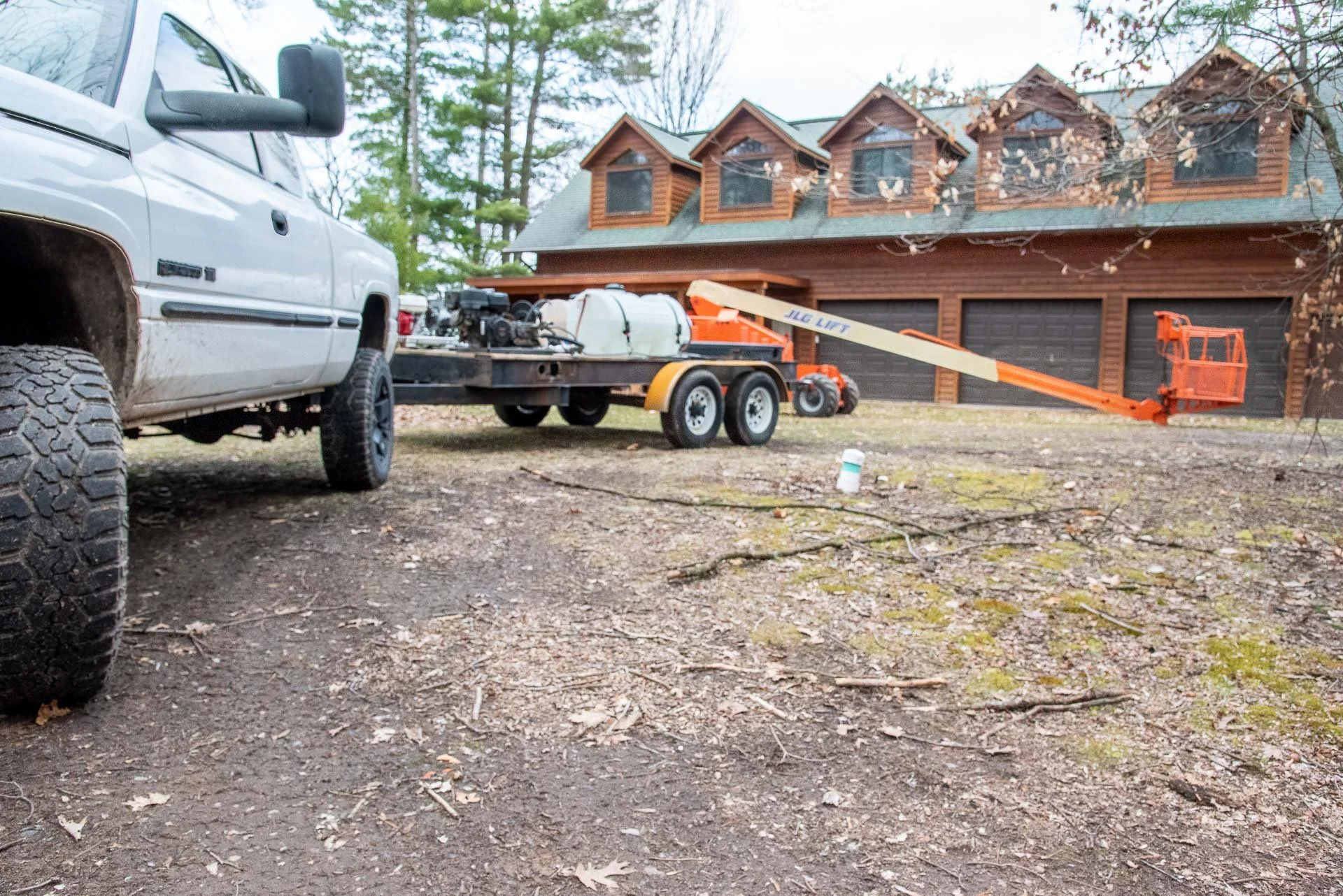 A white pickup truck parked on a dirt driveway in front of a wooden house with a garage. There is a trailer attached to the truck with equipment, including a JLG lift extending out to the right. The house has multiple small dormer windows and brown wooden siding.