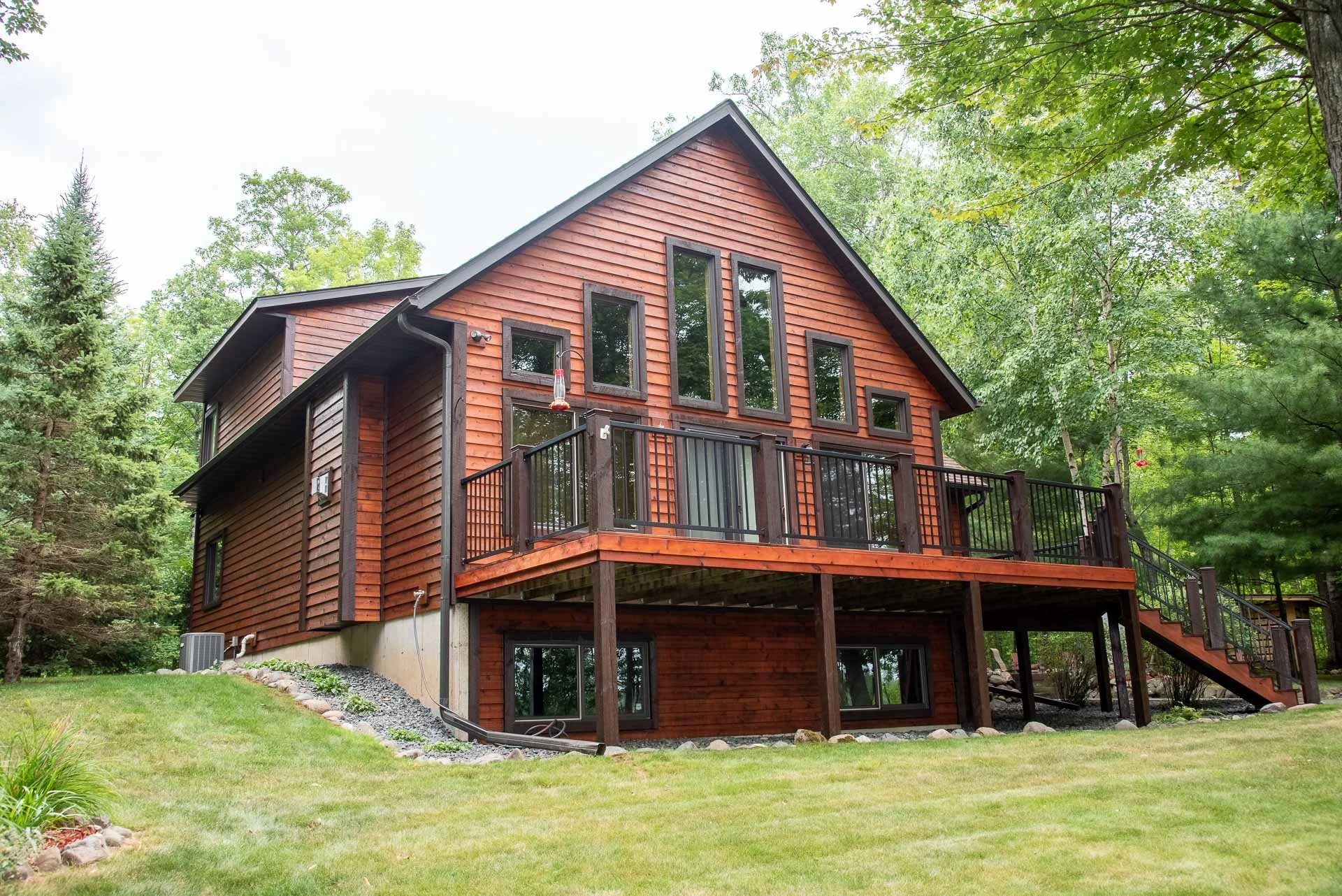 A two-story wooden house with a large upper deck surrounded by green trees and a grassy yard.