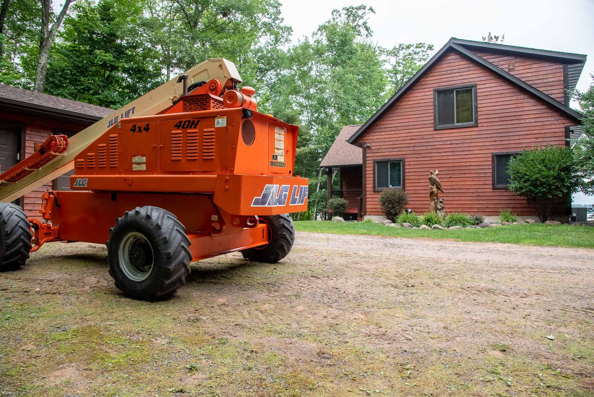 Orange JLG lift with large tires parked on a dirt driveway in front of a brown wood house with a sloped roof, surrounded by green trees and shrubs.