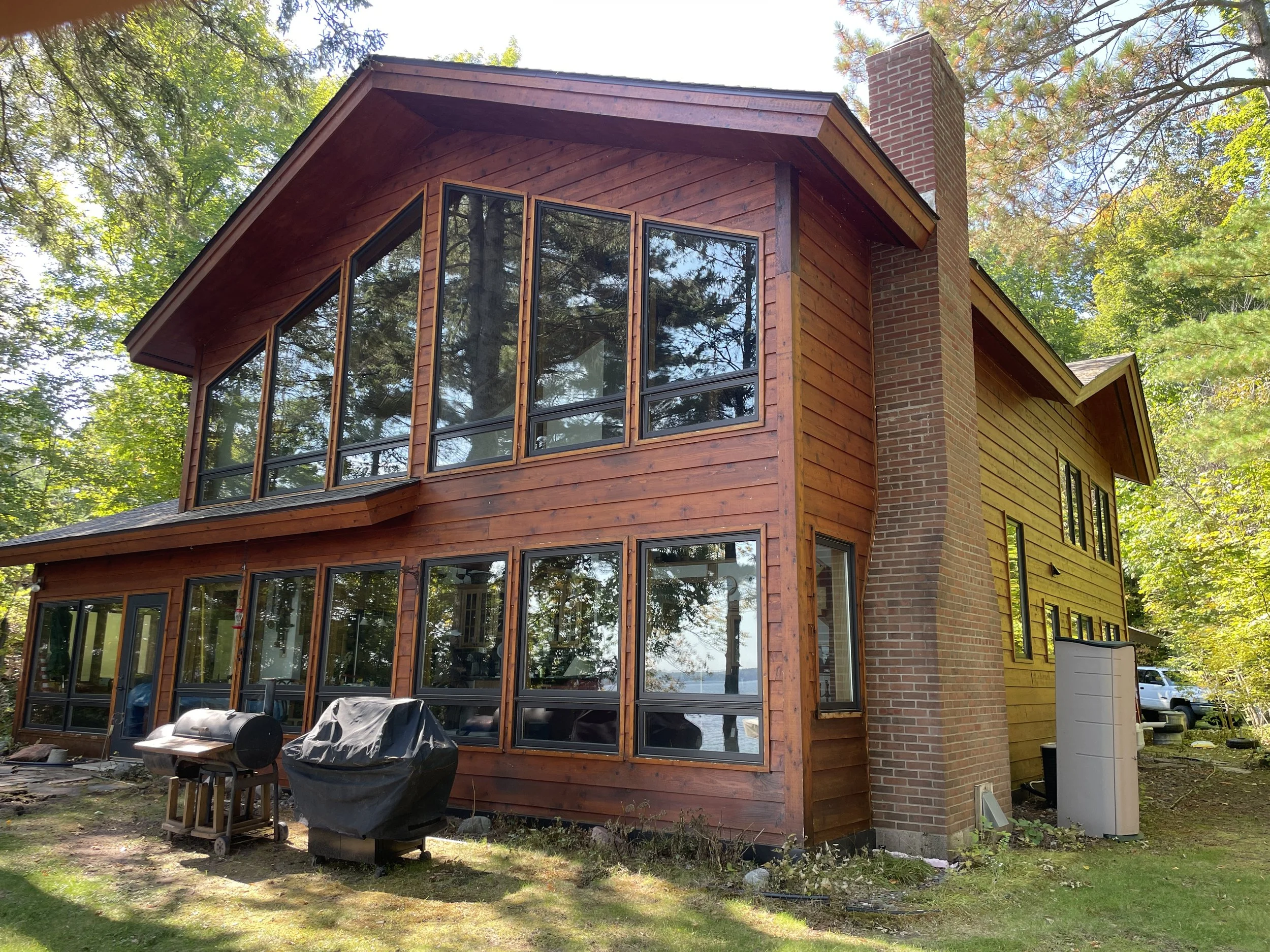 A two-story house with large glass windows, wooden and brick exterior, and surrounded by trees.