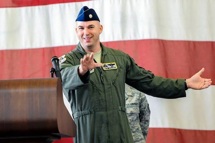 Vanguard 1 Leadership - Jerry Cook in a military uniform with a blue beret, smiling and gesturing with open arms in front of a red, white, and blue backdrop.