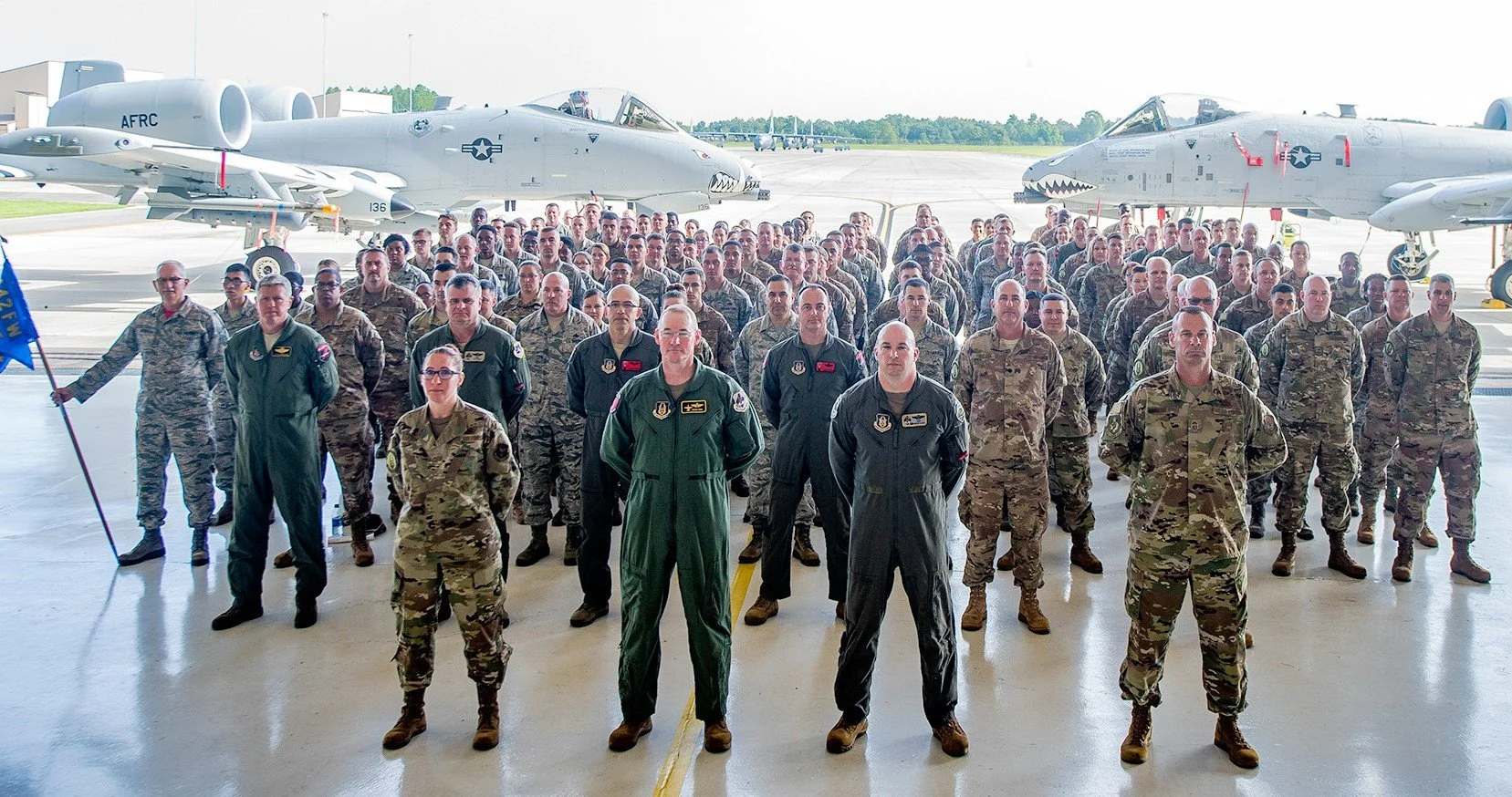 Vanguard 1 Leadership  - Group of military personnel in uniform standing in formation inside an aircraft hangar, with fighter jets parked outside.