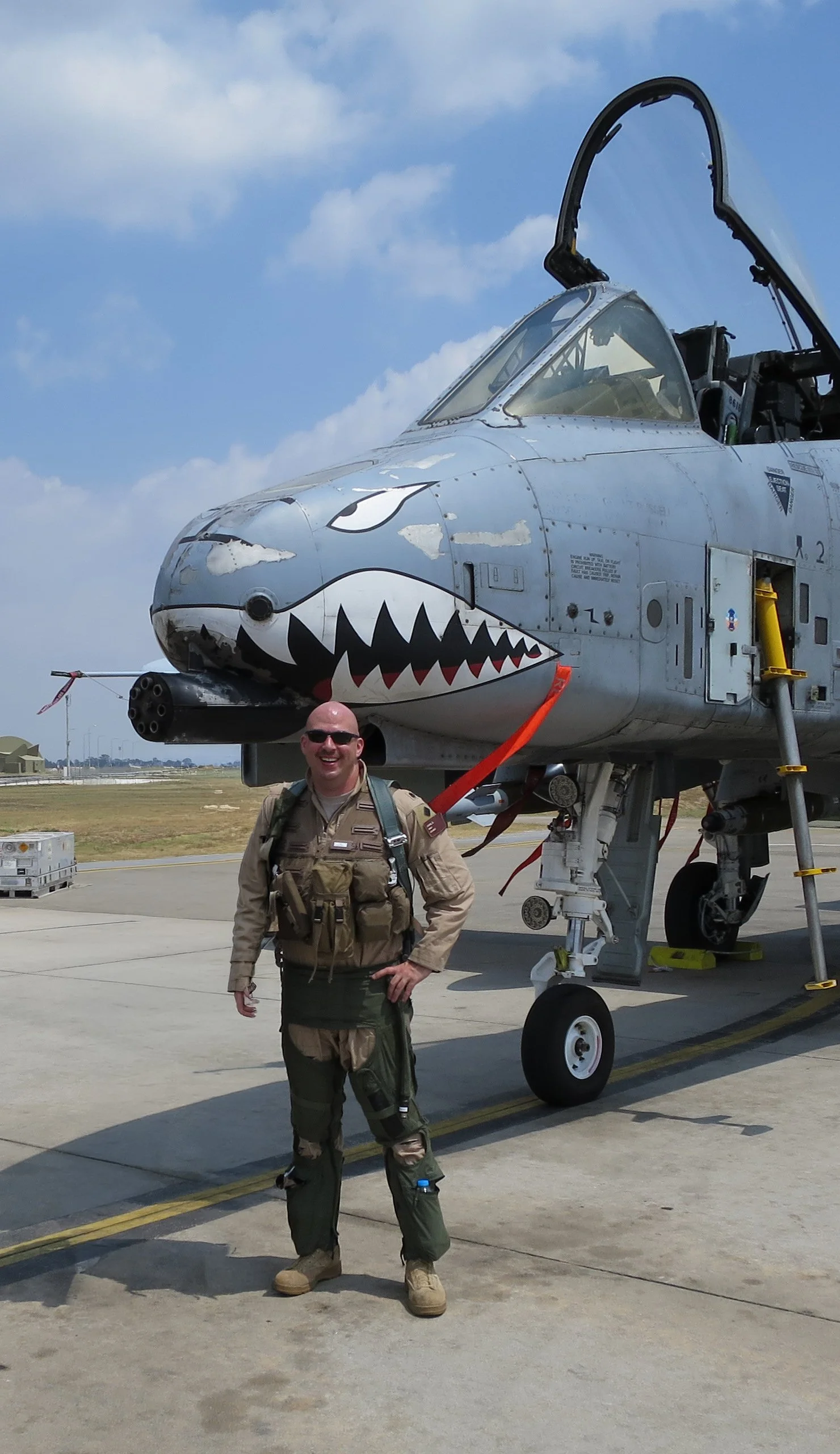 Vanguard 1 Leadership - Jerry Cook in flight gear standing in front of a fighter jet with a shark face painted on its nose, on an airport tarmac with a blue sky background.