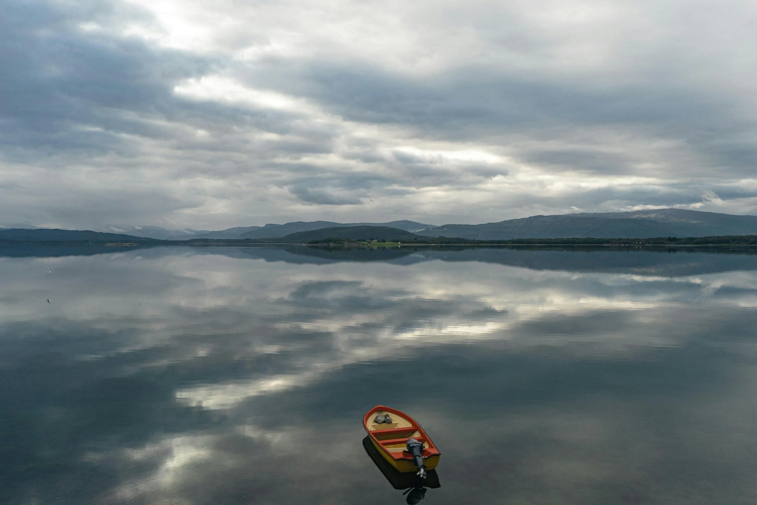A small orange rowboat floating on a calm lake, with gray clouds overhead and mountains in the distance, reflected in the water.