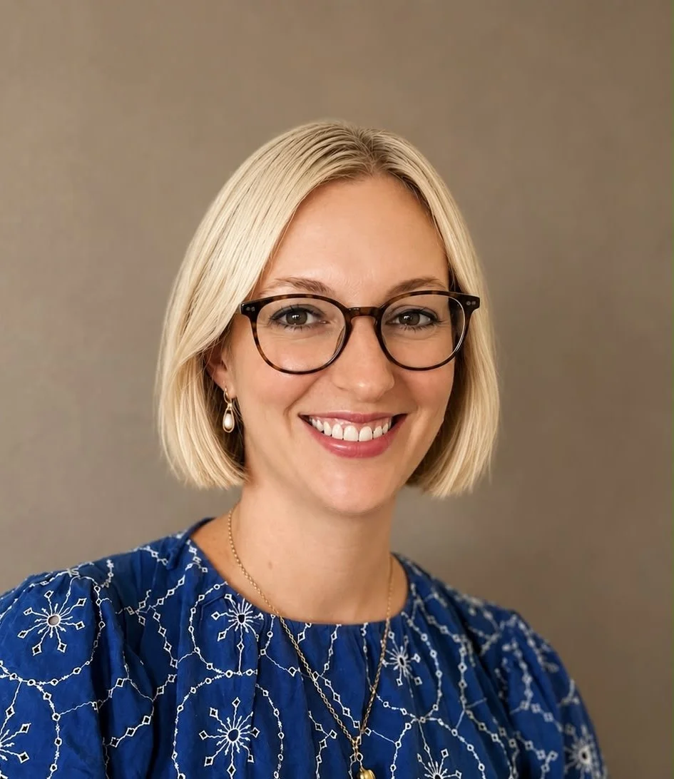 A woman with blonde hair, glasses, and earrings smiling at the camera. She's wearing a blue embroidered blouse and layered necklaces, standing against a neutral background.