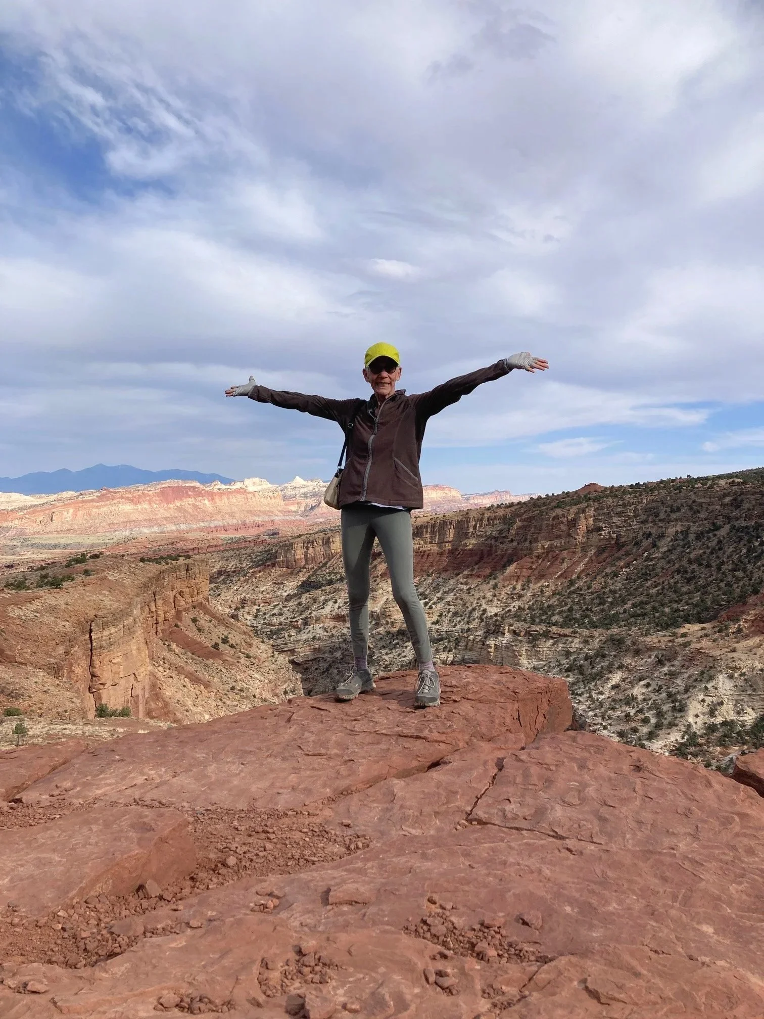 A woman standing on a large rock ledge in a canyon, with arms outstretched, wearing a yellow cap, sunglasses, a dark jacket, gray leggings, and hiking shoes, with vast canyon landscape and cloudy sky in the background.