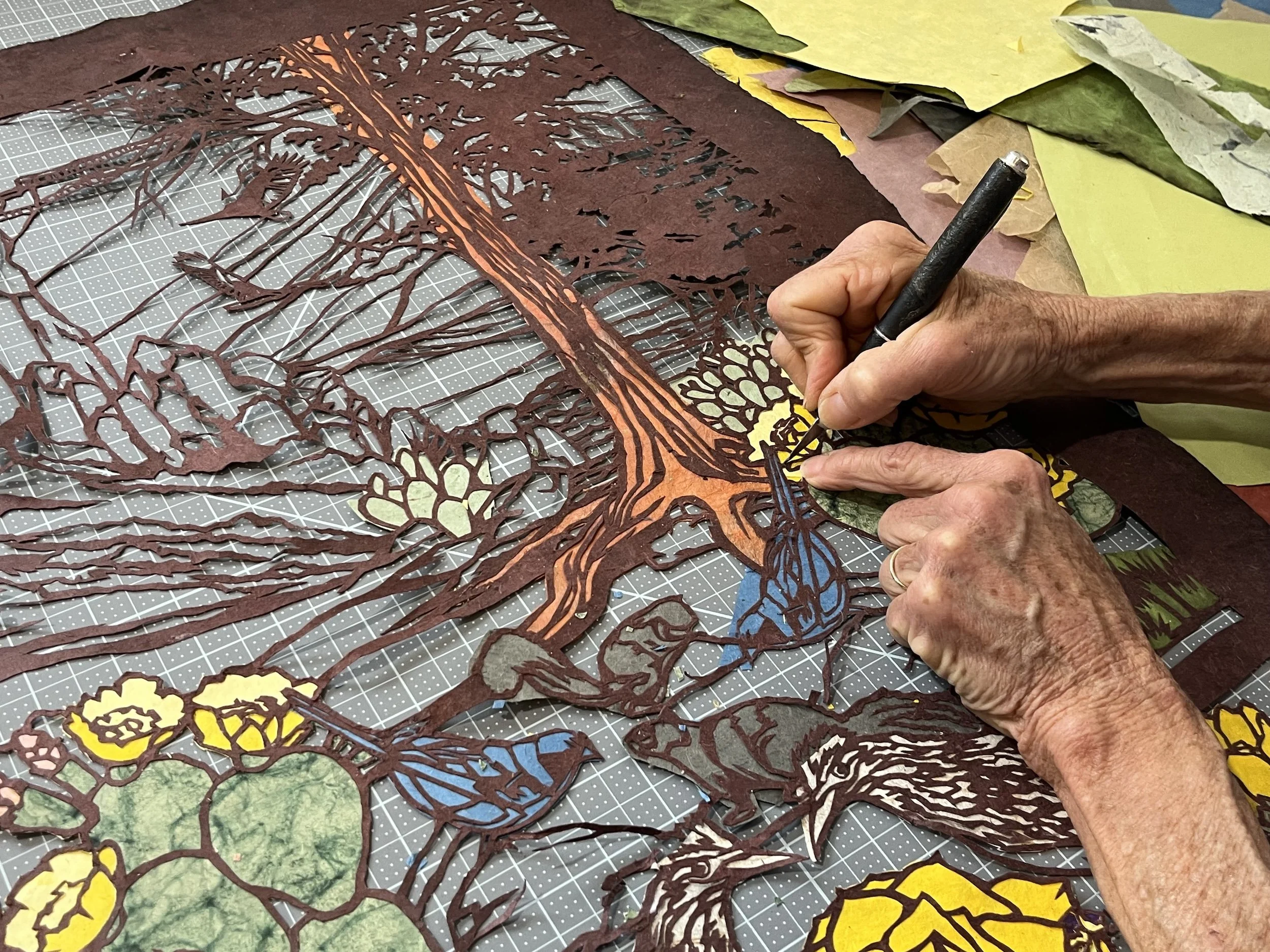 Person creating a paper cutout artwork featuring a large tree, flowers, birds, and a squirrel, with various colored paper sheets surrounding the workspace.
