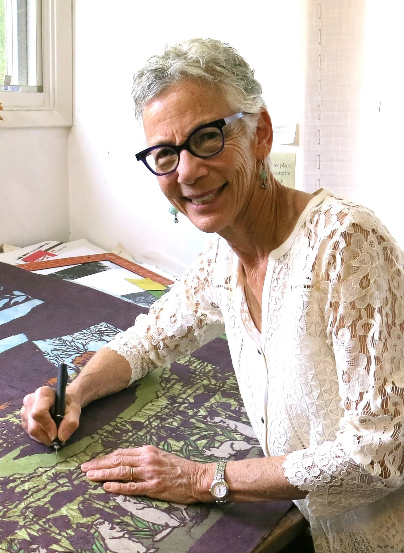 A woman with short gray hair, wearing glasses, earrings, a lace white blouse, and a watch, is sitting at a table signing artwork with a pen, smiling at the camera inside an art studio.