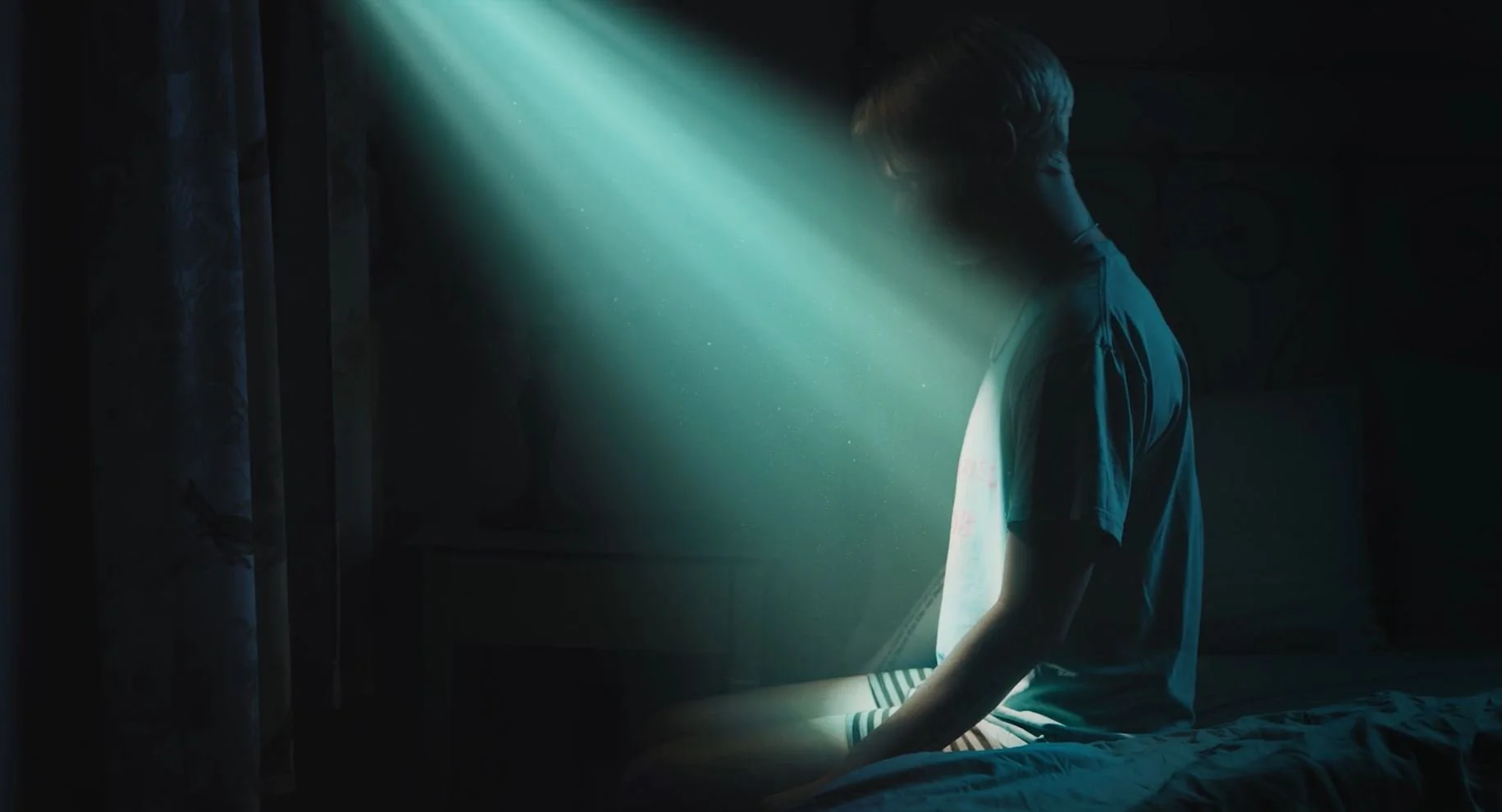 A young person sitting on a bed in a dimly lit room, with sunlight streaming through a window and illuminating part of their face and body.