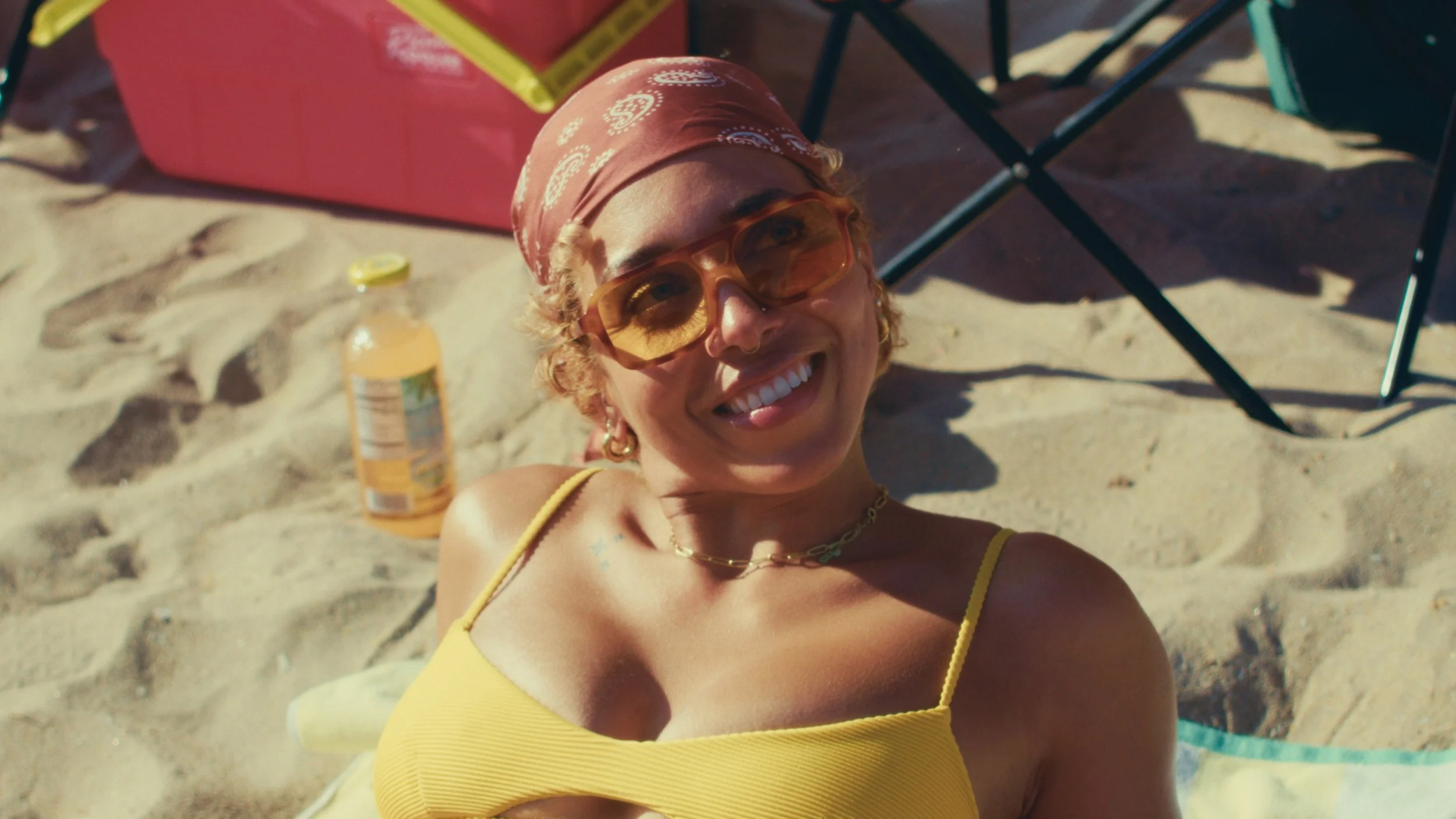 A woman lying on the sand at the beach, smiling, wearing yellow sunglasses and a yellow tank top, with a pink cooler and chairs in the background.