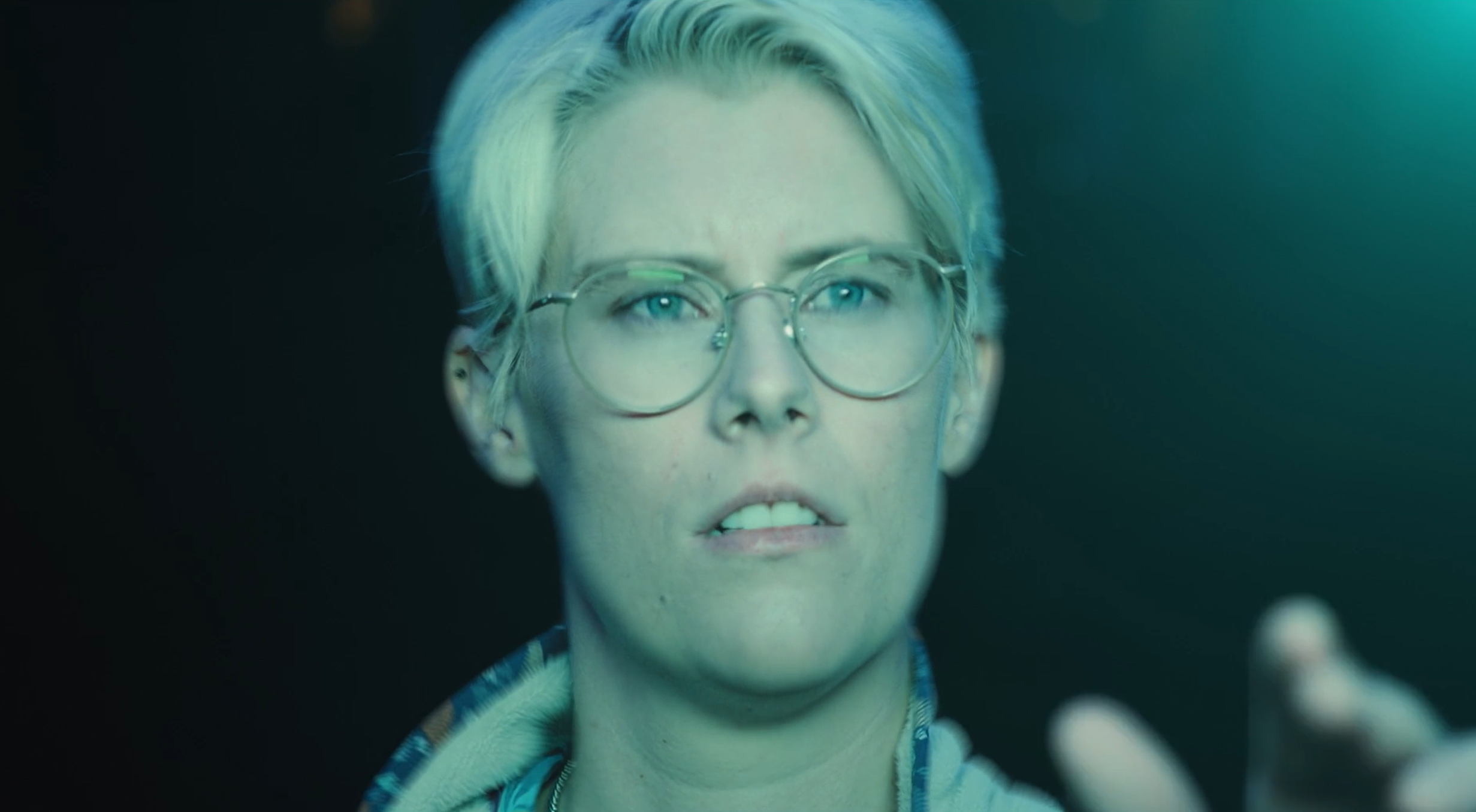 Close-up shot of a woman with short blonde hair and large glasses, reaching out with her hand, against a dark background