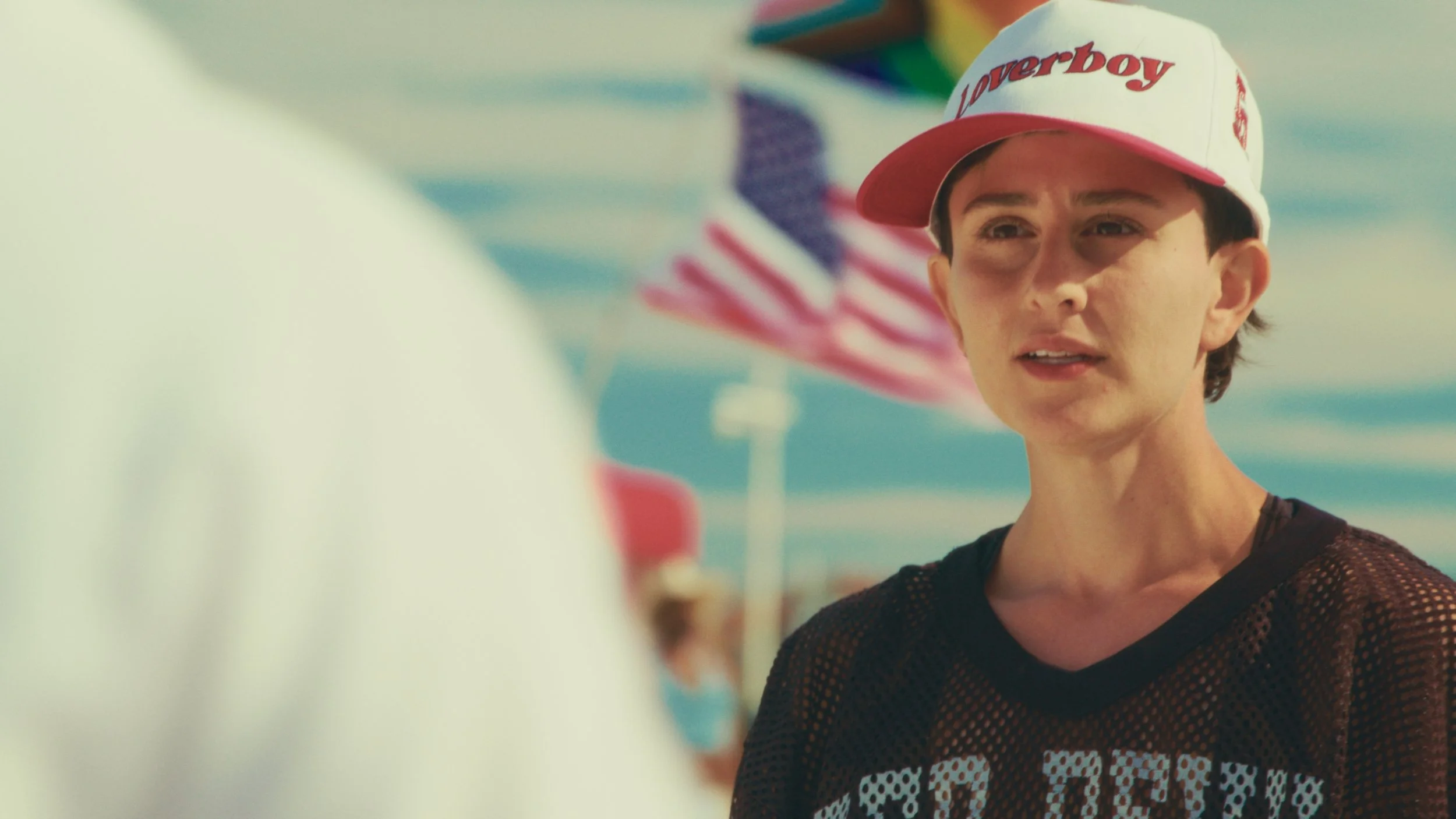 A woman wearing a red and white 'Loverboy' baseball cap and black mesh shirt talking to someone against a backdrop of an American flag and a beach scene.