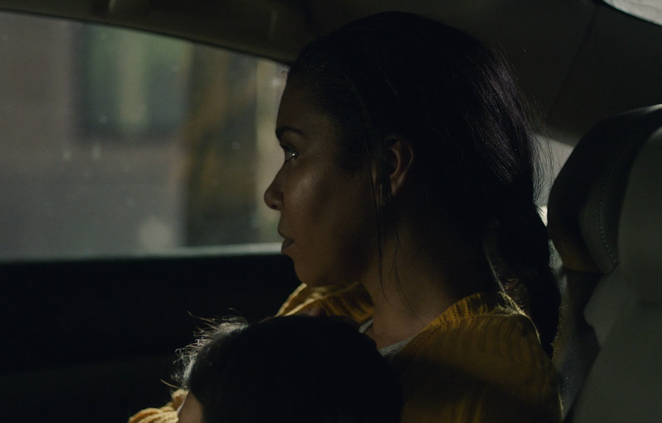 A woman with dark hair sitting in a car, looking ahead, with a child resting on her lap.