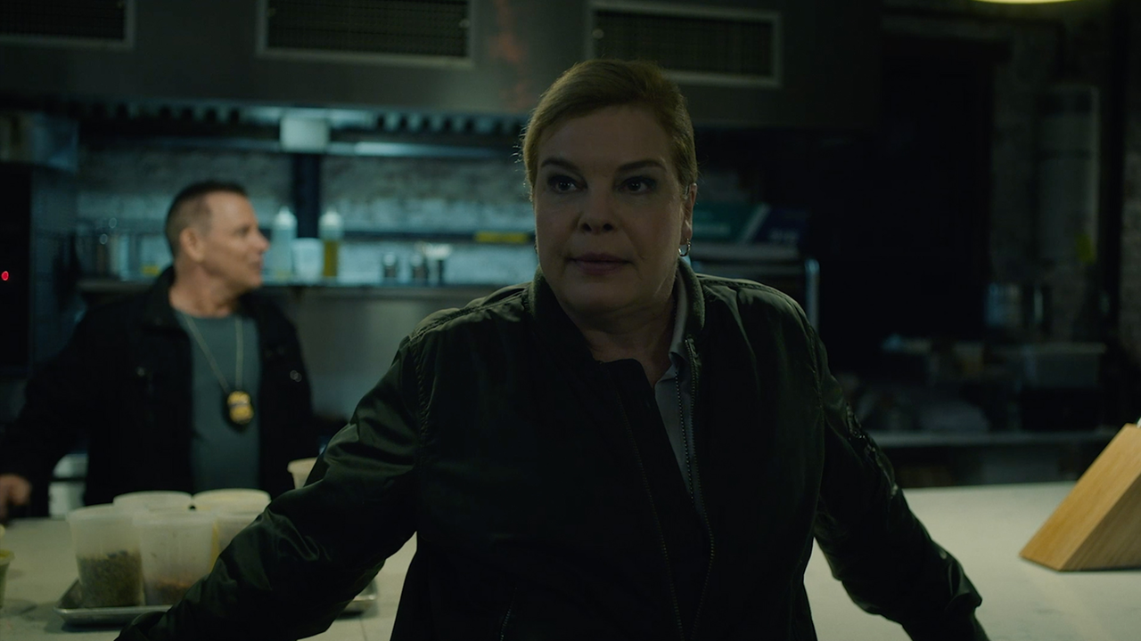 Catherine Curtin in a dark jacket in a kitchen or warehouse setting, with a man with a badge in the background.