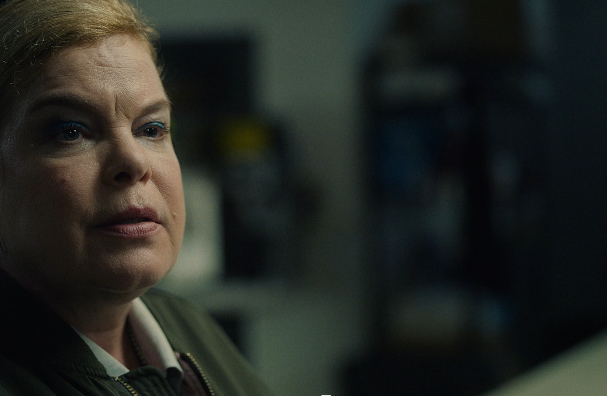 Close-up of a woman with short, blond hair, wearing makeup with blue eyeshadow, looking to the side with a serious expression in a dimly lit room.