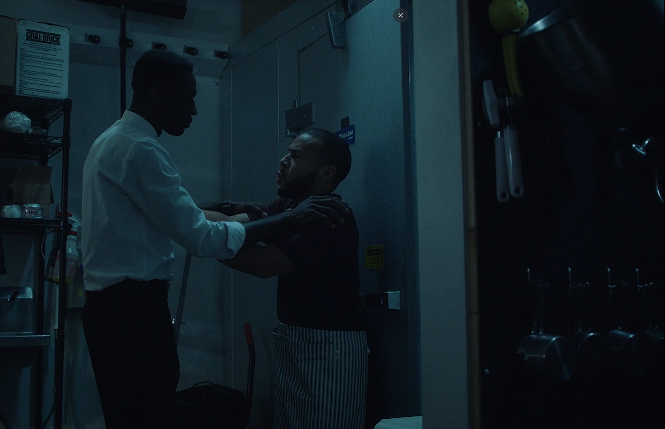 A person wearing a white shirt helping another person with a beard, dressed in a black shirt and striped pants, in a dimly lit kitchen.