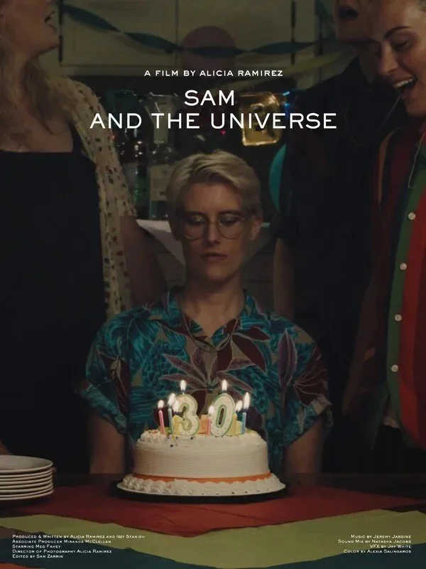 A woman with short blonde hair and glasses is sitting at a table with a birthday cake and 13 candles. She is wearing a colorful blouse and appears to be at a birthday celebration. Two women are standing beside her, one on each side, and there are sta
