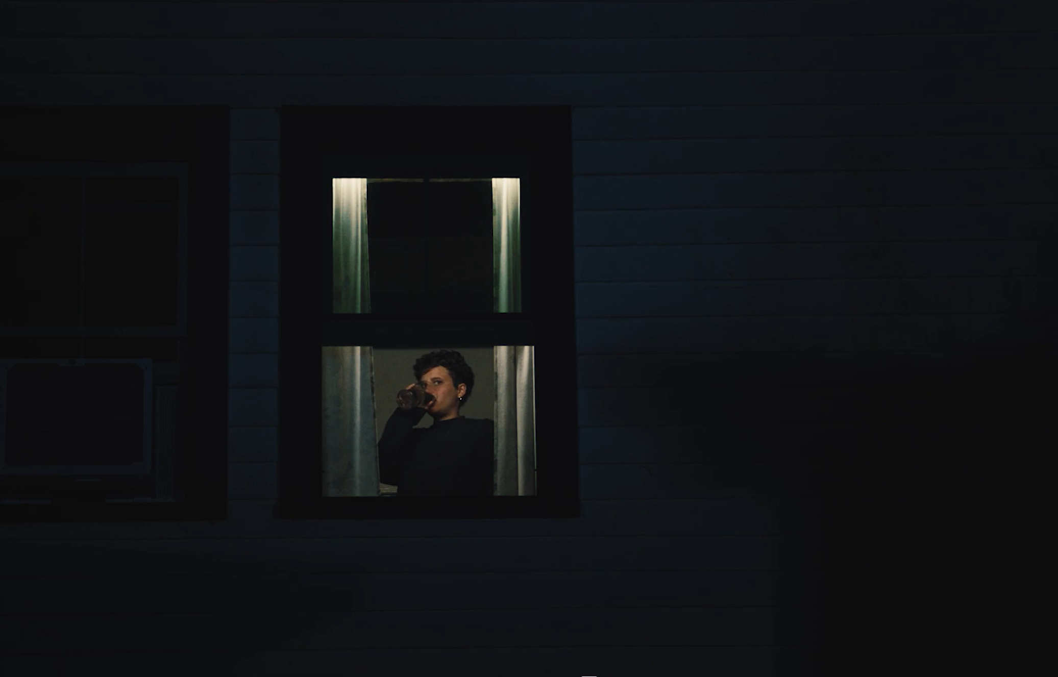 A person with short dark hair drinking from a bottle through a window at night.
