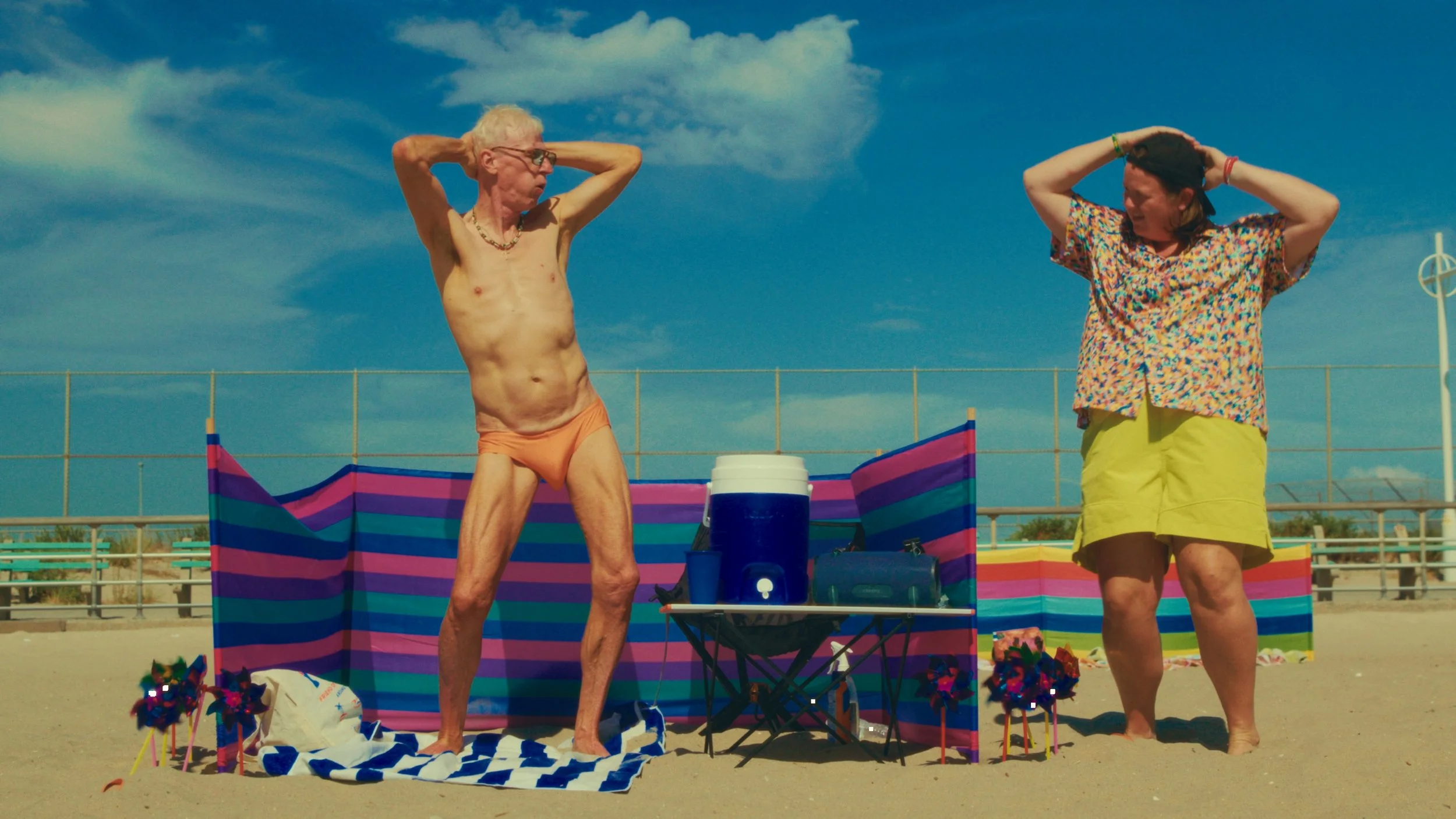 An elderly man and a woman standing on a beach, wearing summer clothing, with beach chairs, and a colorful striped beach backdrop in the background.
