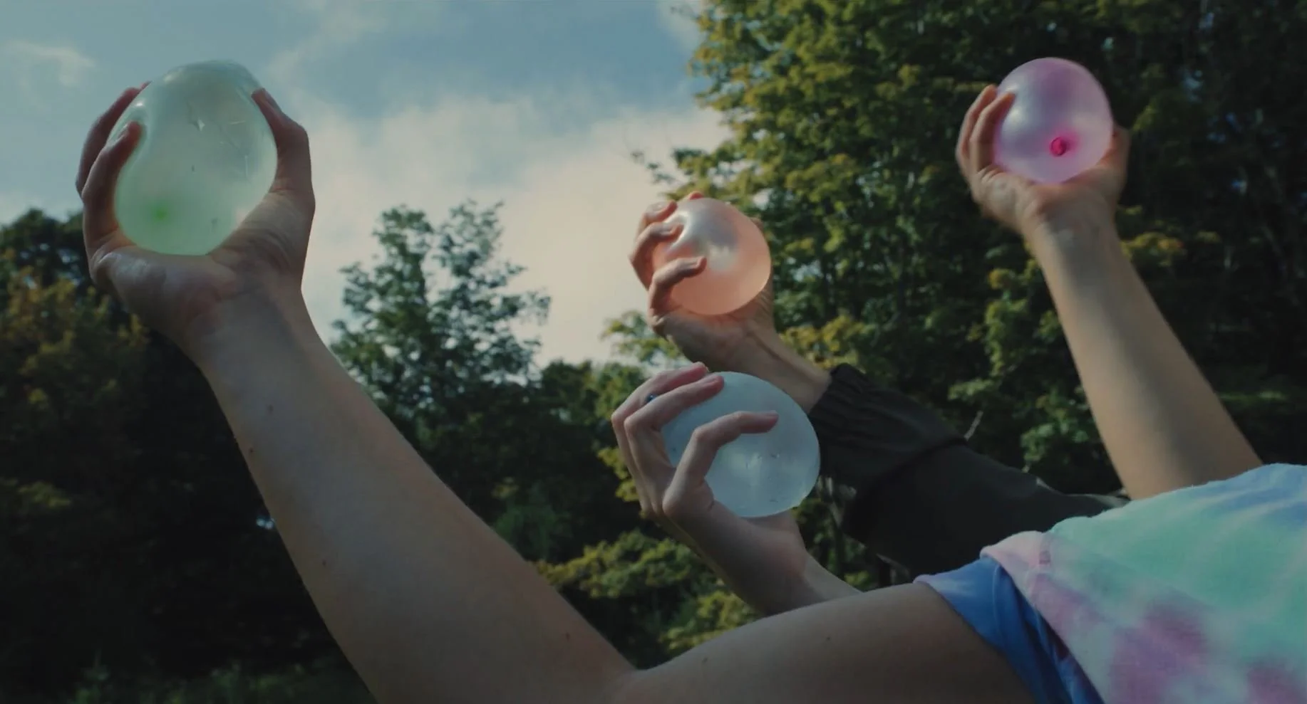 Four people lying on the ground outdoors, each holding a colorful, translucent bubble ball up in the air with one hand. The background shows trees and a partly cloudy sky.