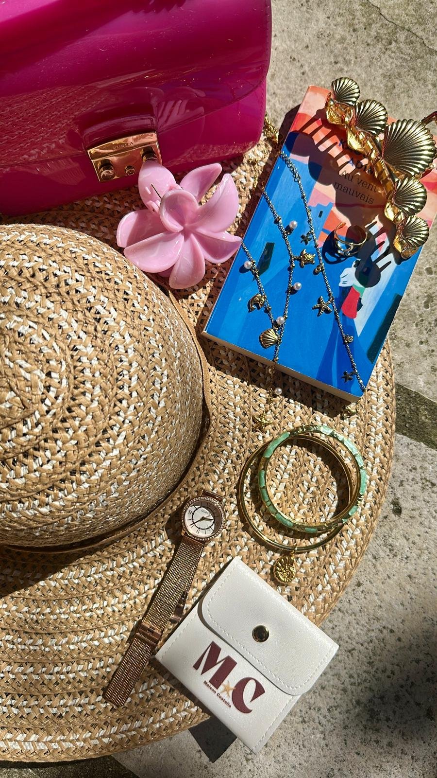 Accessoires de mode et objets de décoration sur un tapis en osier, comprenant un chapeau en rotin, un bracelet doré, une montre, des colliers avec pendentifs, un portefeuille blanc avec inscription, un gros bracelet vert et or, des bijoux dorés avec des perles, une bague, une bande de papier avec un message, un sac rose en plastique, et un bijou en forme de fleurs dorées.