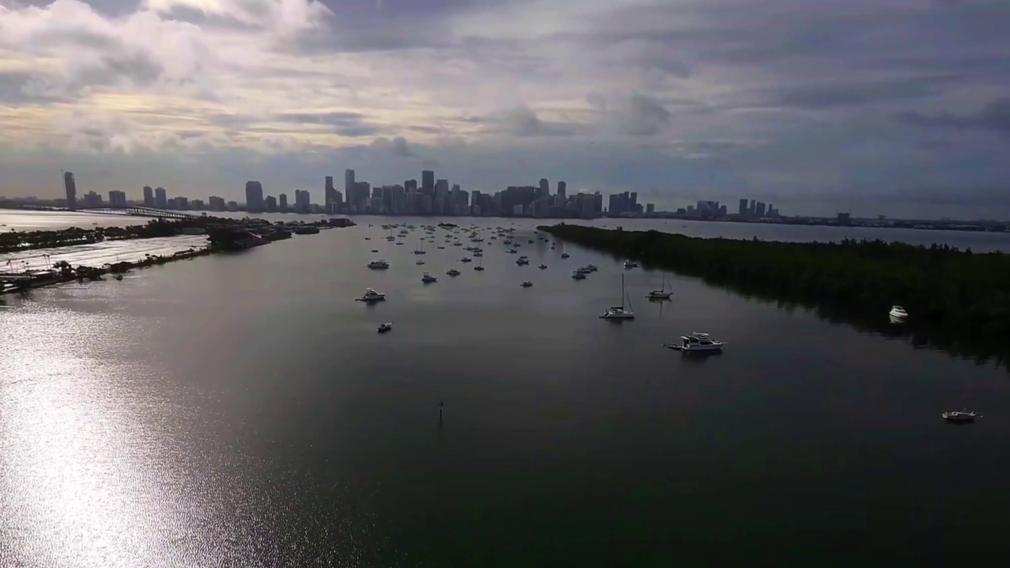 Aerial view of a city skyline across a body of water with numerous boats and yachts anchored, surrounded by green land, under a cloudy sky.
