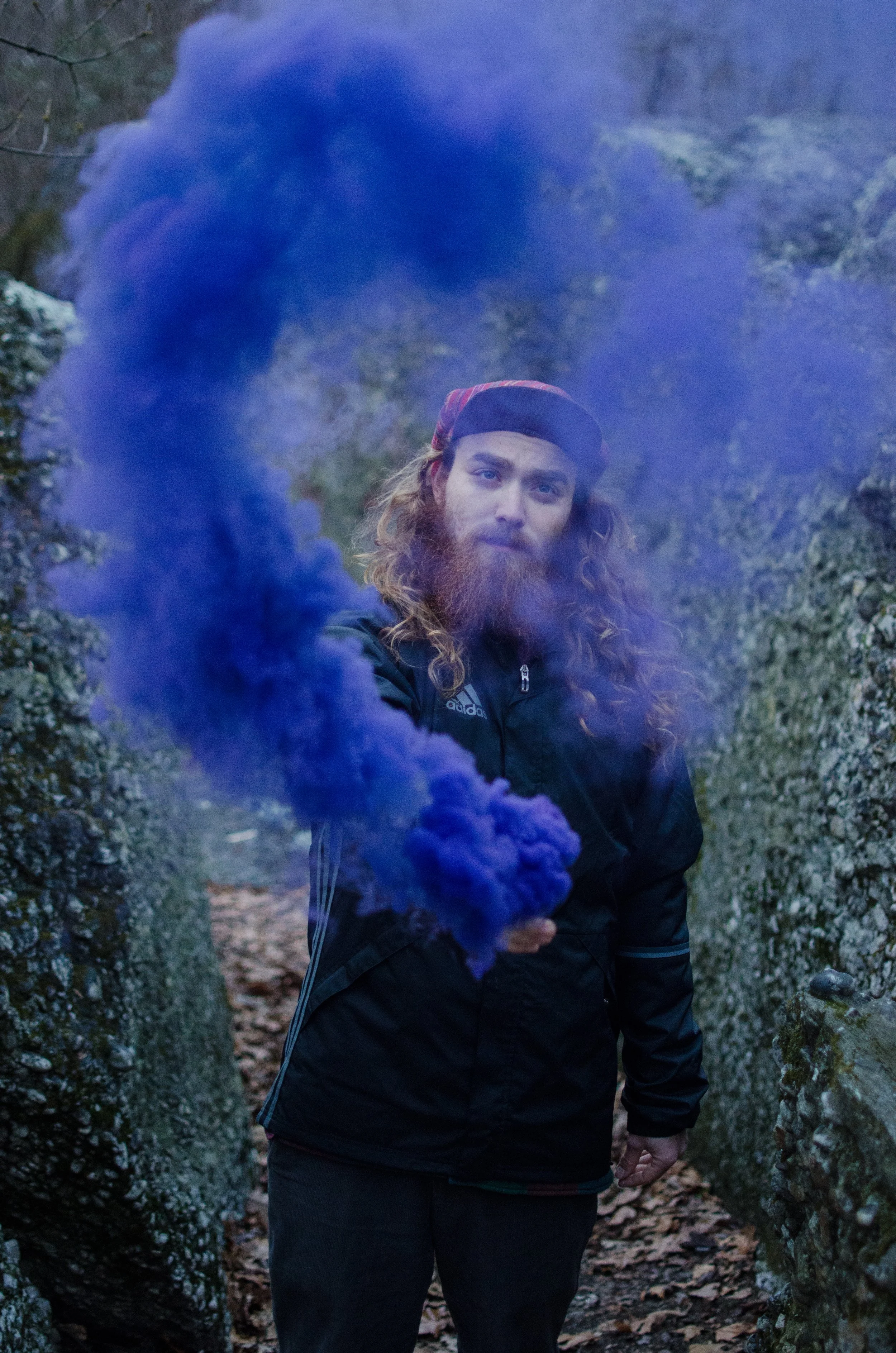 A man with long wavy hair and a beard, wearing a black jacket and a backwards cap, stands between moss-covered rocks on a forested trail while holding a blue smoke grenade that emits a dense cloud of blue smoke.