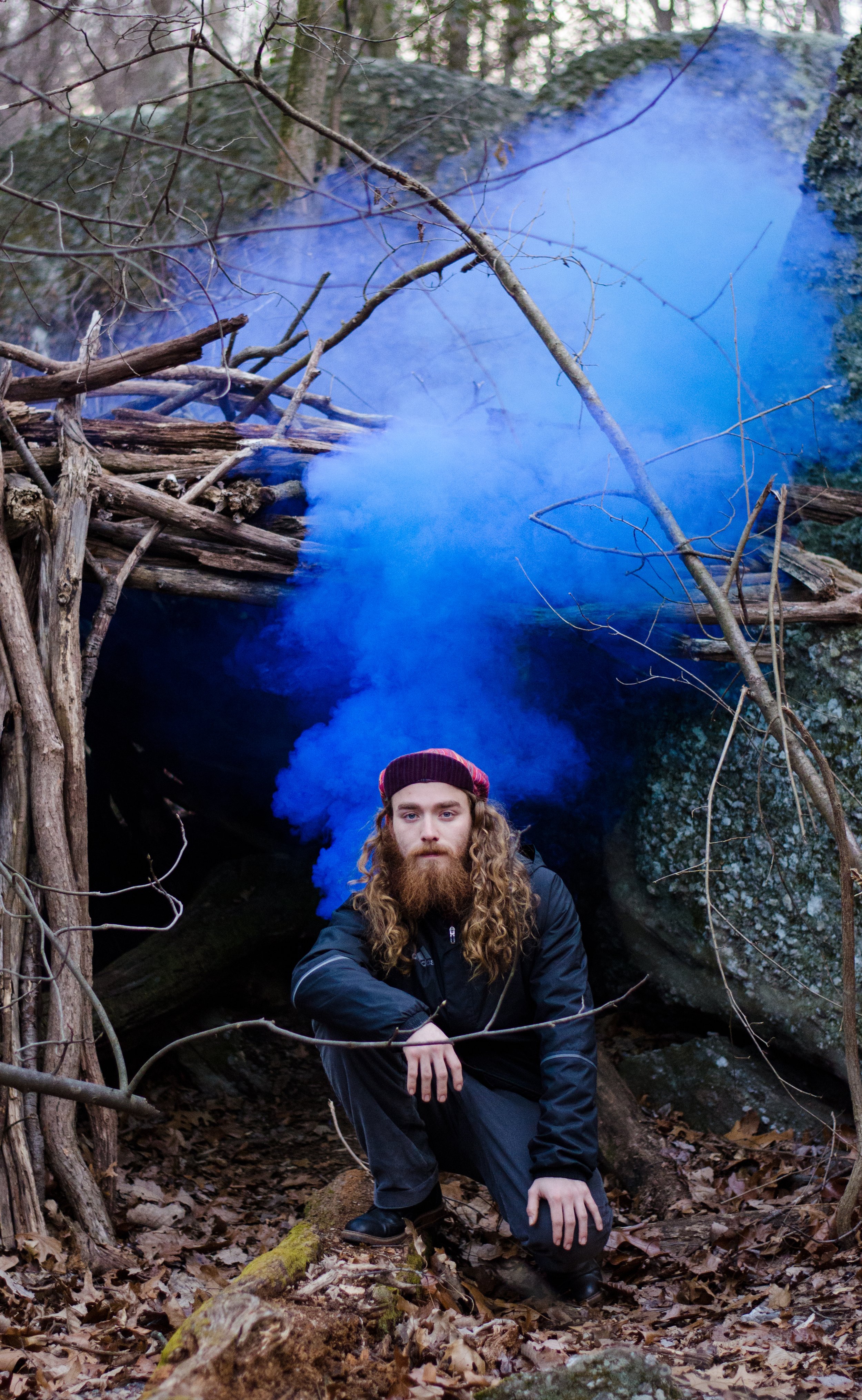 A man with long hair and a beard crouching in a forest, surrounded by fallen leaves and leafless branches, with a mist of blue smoke behind him and a rocky background.