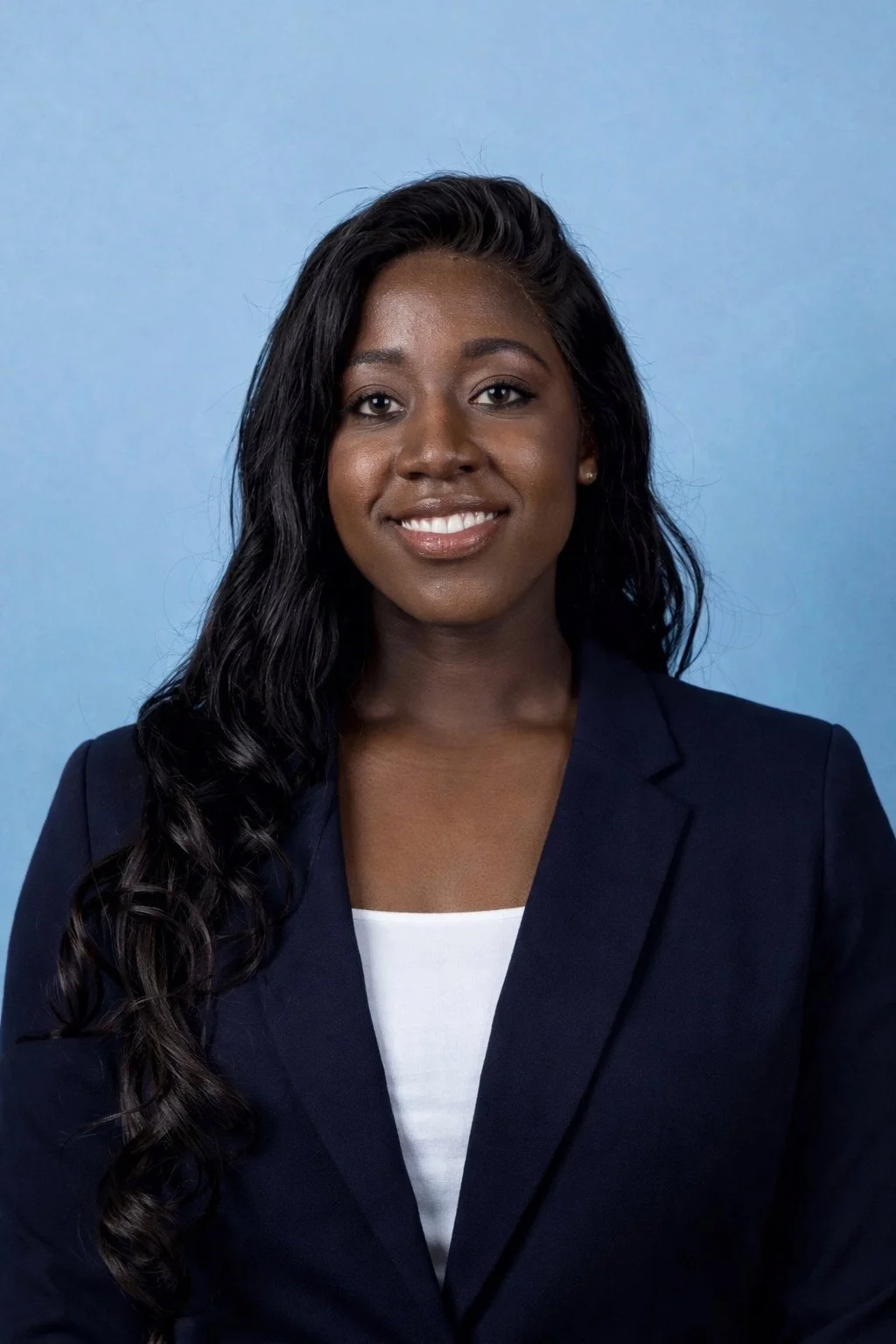 A professional woman with long, wavy black hair, wearing a navy blazer over a white top, smiling against a light blue background.