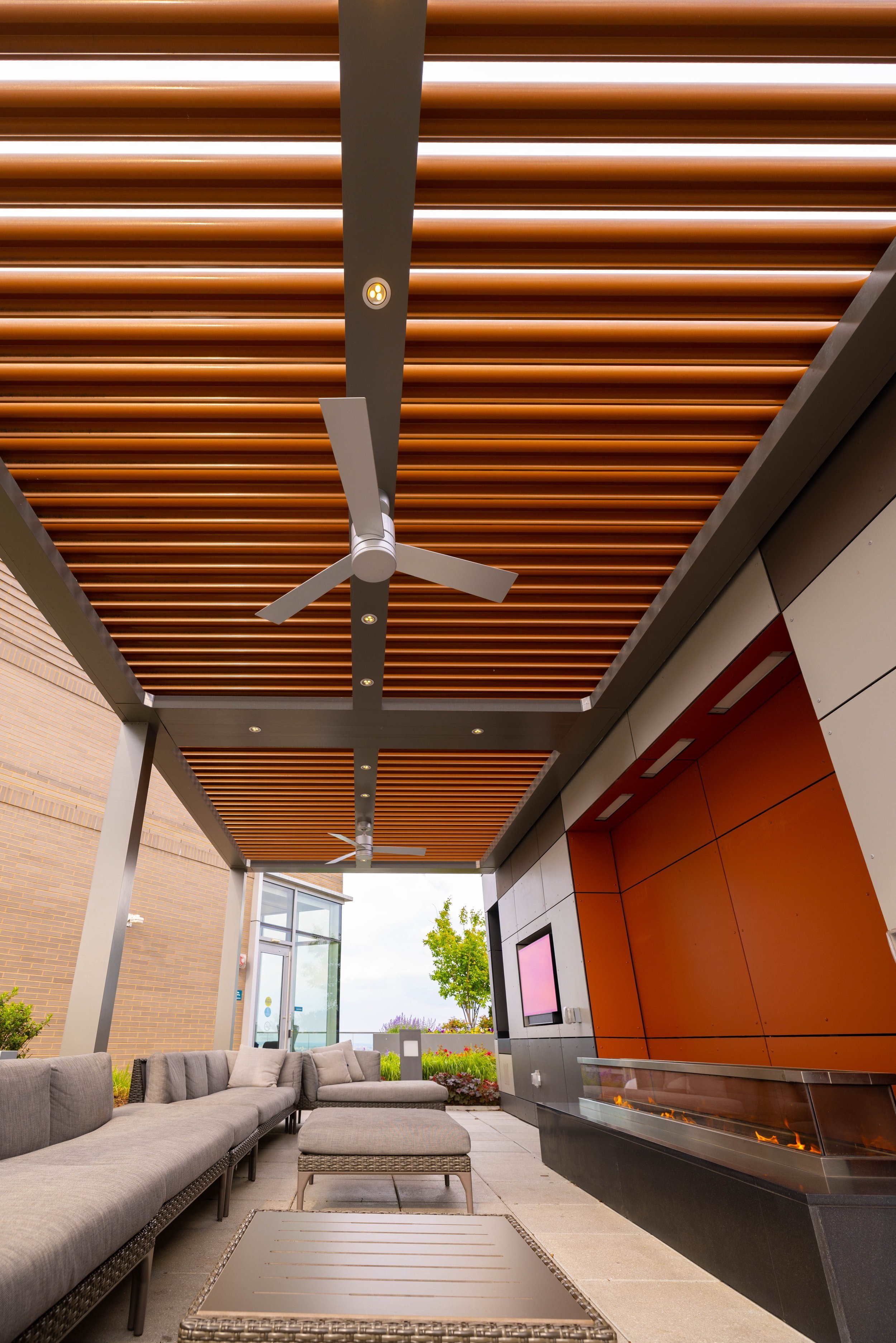Outdoor seating area with a long gray sectional sofa, a matching ottoman, and a rectangular metal table. The ceiling has wooden slats and recessed lighting, and there are two ceiling fans. A gas fireplace with flames is built into the wall, and outdoor plants and trees are visible beyond the covered patio.