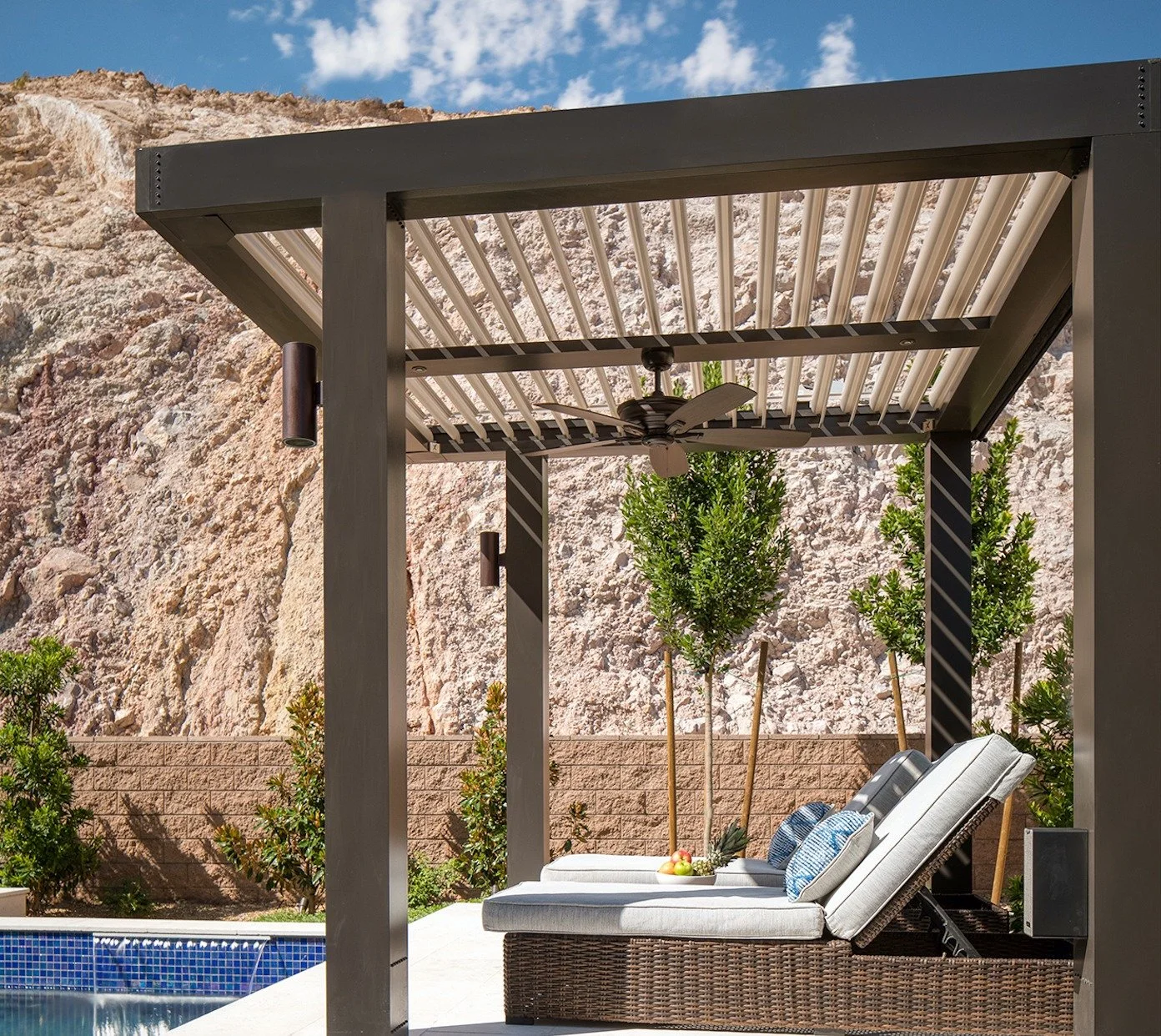 Backyard pool area with a pergola, lounge chairs, a ceiling fan, and a stone wall in the background.