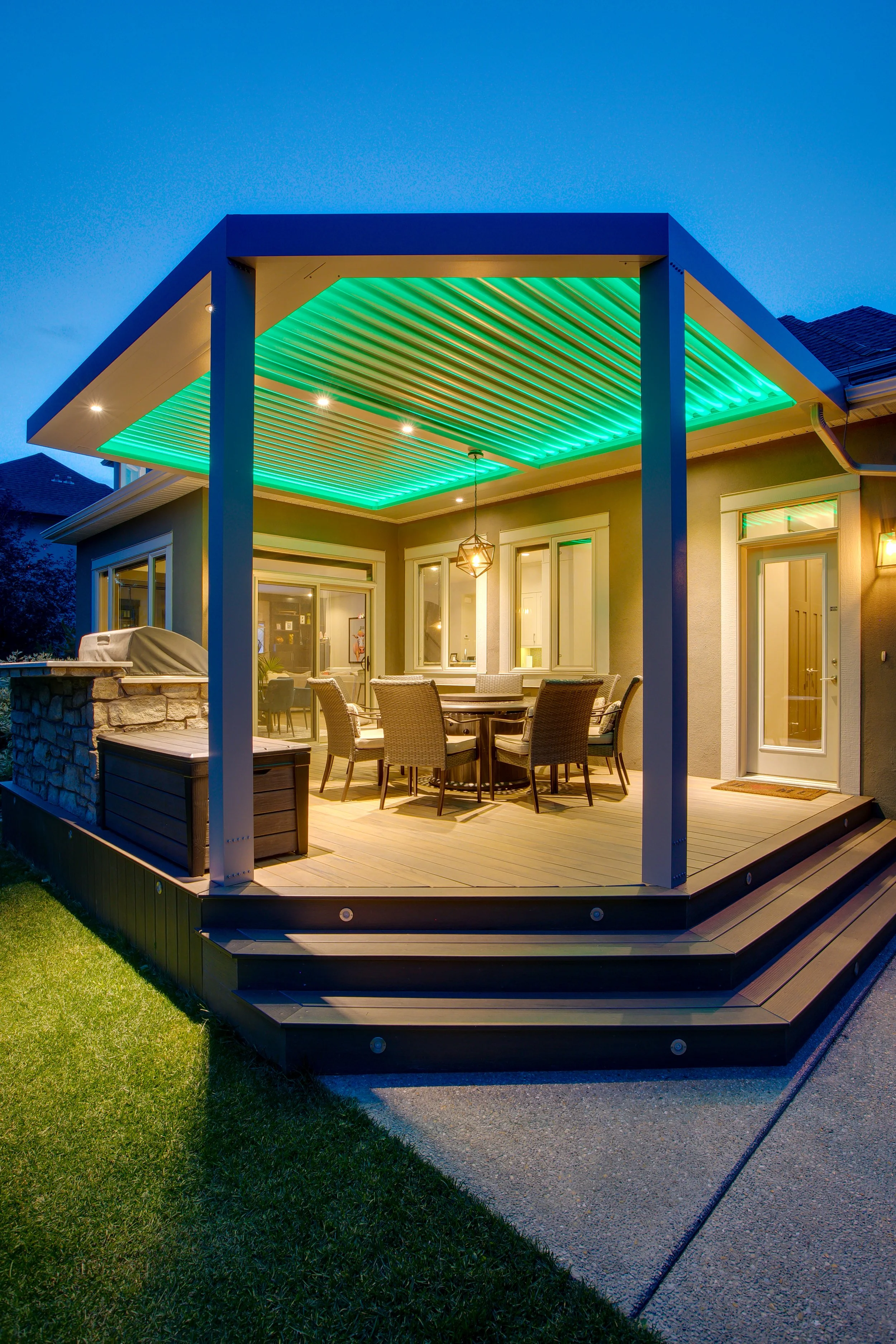 Backyard patio with wooden deck, outdoor dining furniture, and a ceiling with green lighting, attached to a house during early evening.