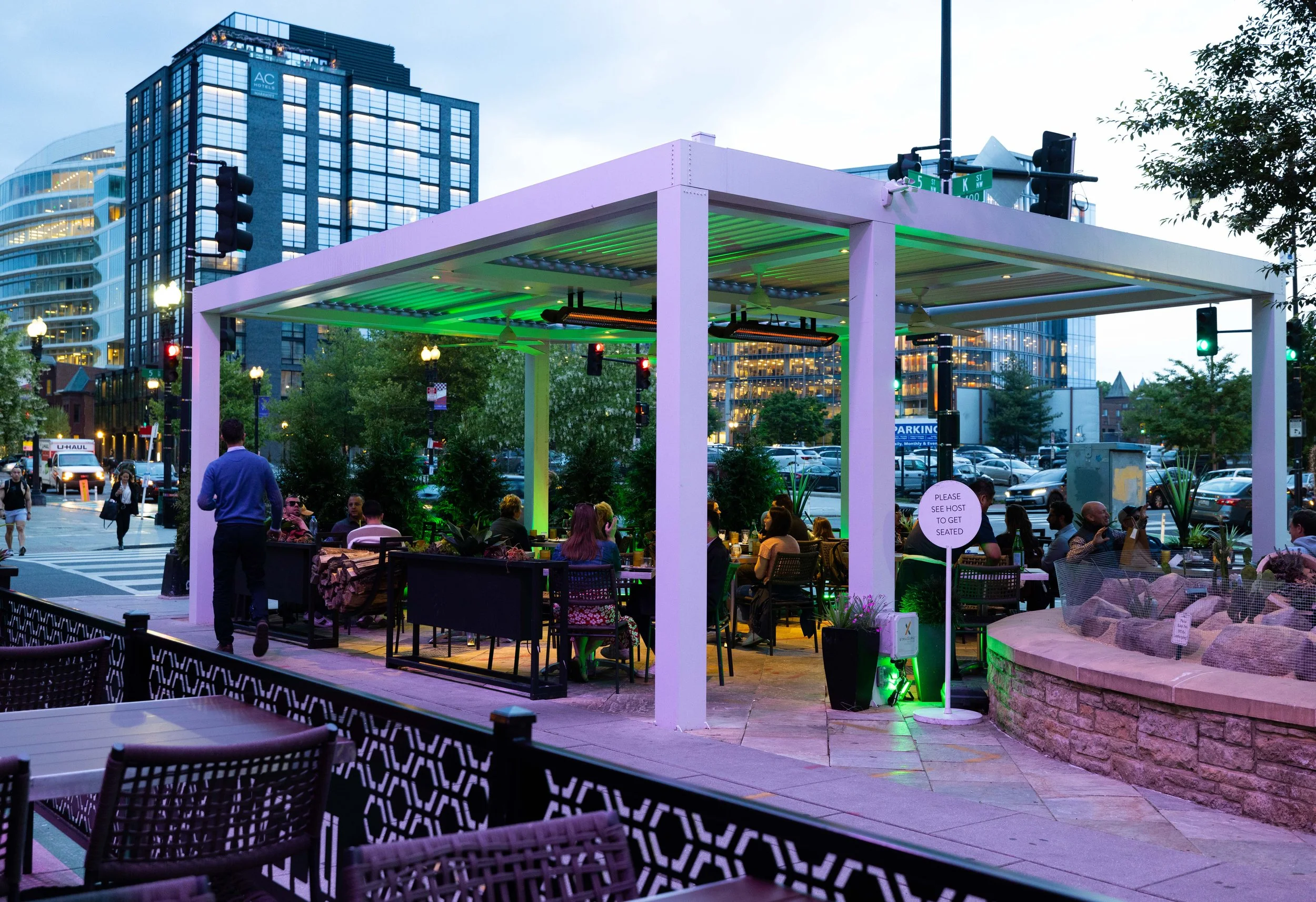 Outdoor restaurant with people dining under a white pergola, city buildings in the background, some illuminated green lights, and a sign to see the host for seating.