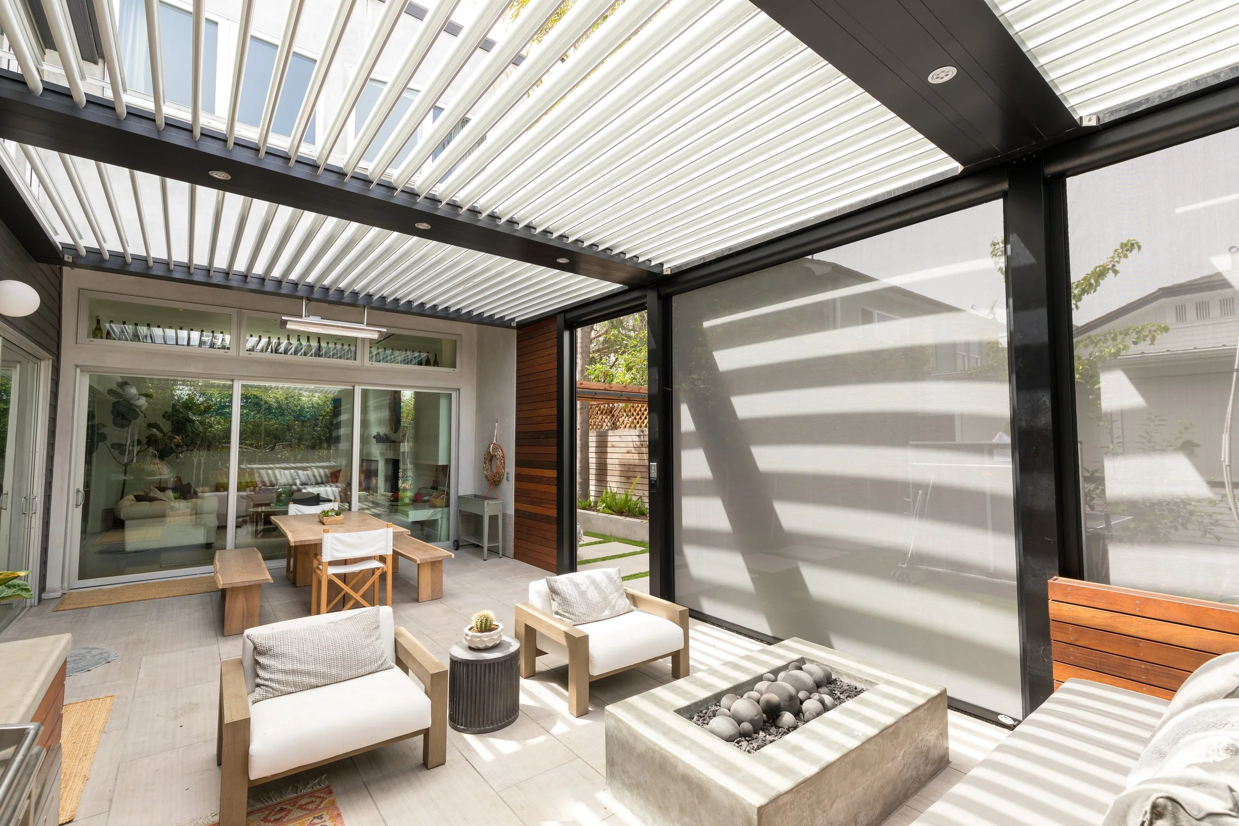 Sunny patio with modern outdoor furniture, a fire pit, and a view into the house's interior through sliding glass doors. The space features a mix of wood, metal, and neutral-colored cushions, with a partially shaded roof.