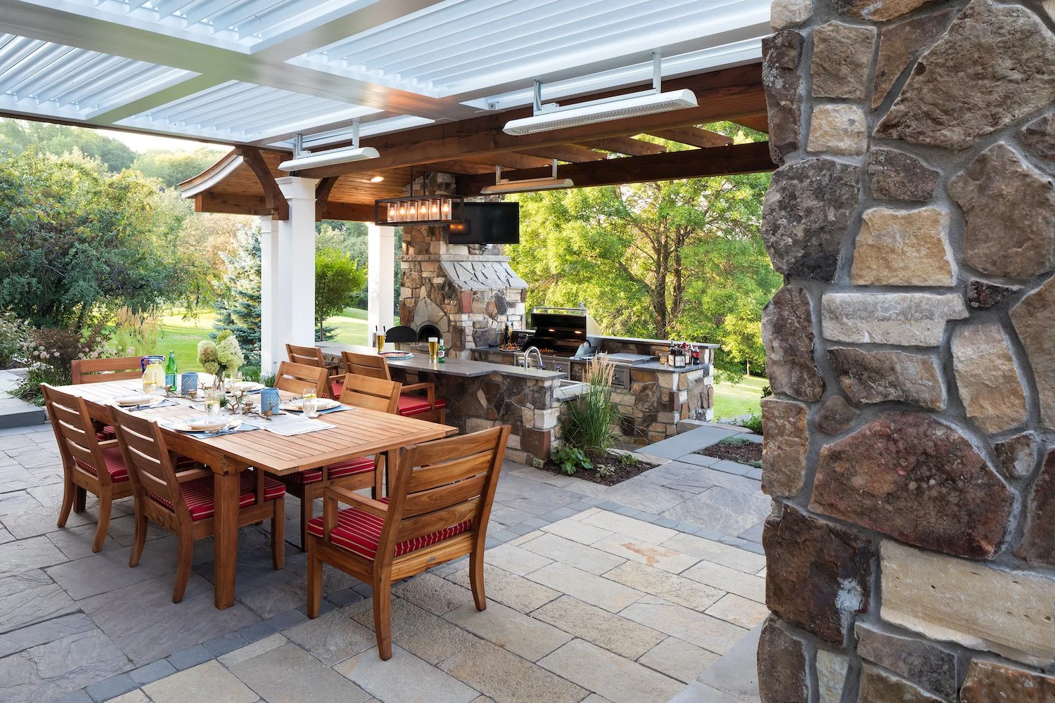 Outdoor patio with dining table set for a meal, with chairs featuring red striped cushions, a stone fireplace, BBQ grill, and a TV mounted on the wooden beam, surrounded by green trees