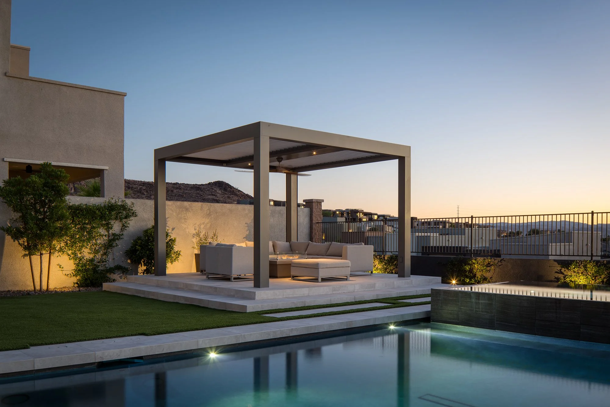 A modern backyard patio area with outdoor furniture, a swimming pool, and landscaping at sunset.