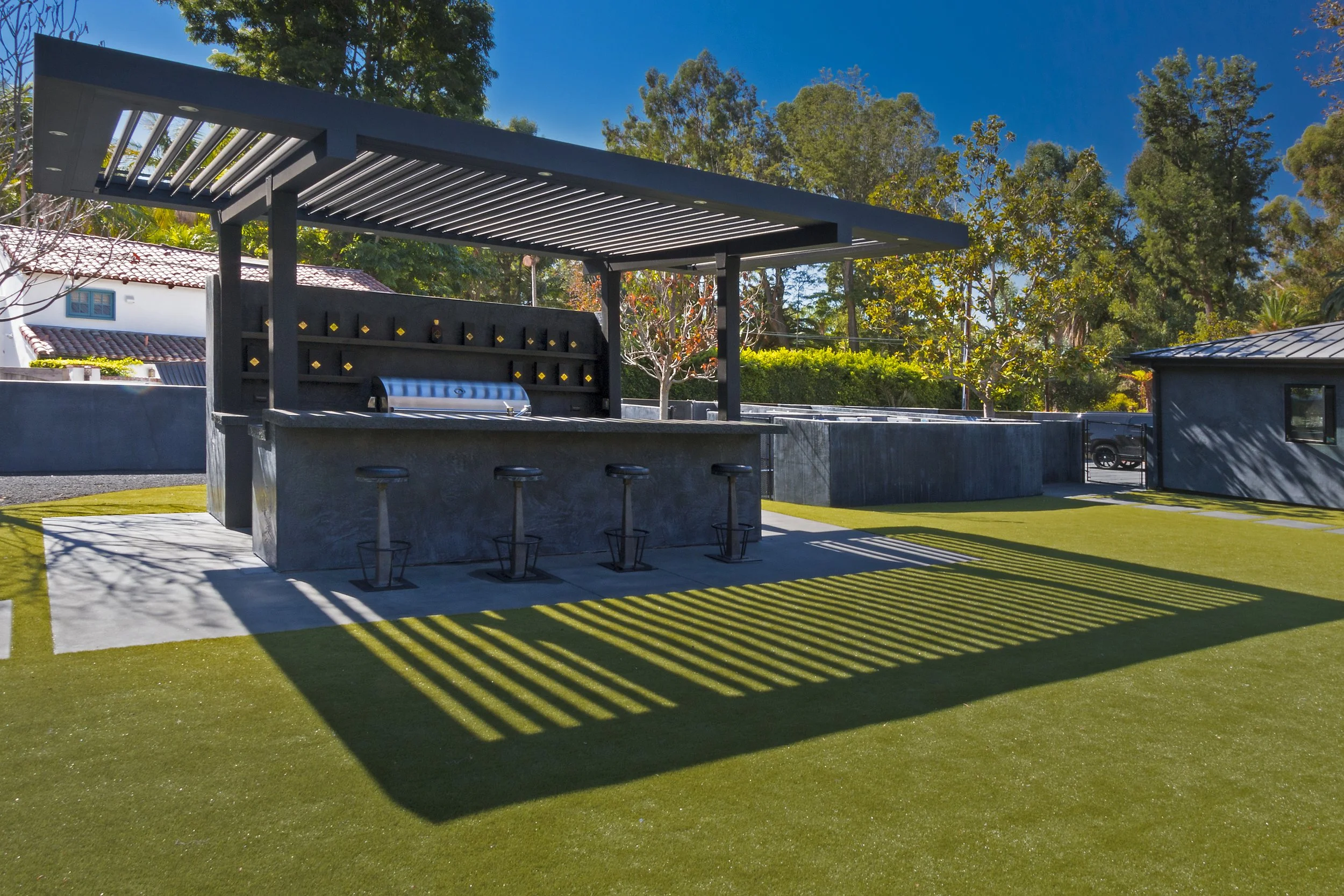 Outdoor bar area with four bar stools, a built-in grill, and a modern shaded structure. The area is surrounded by a green lawn and trees, with a blue sky in the background.
