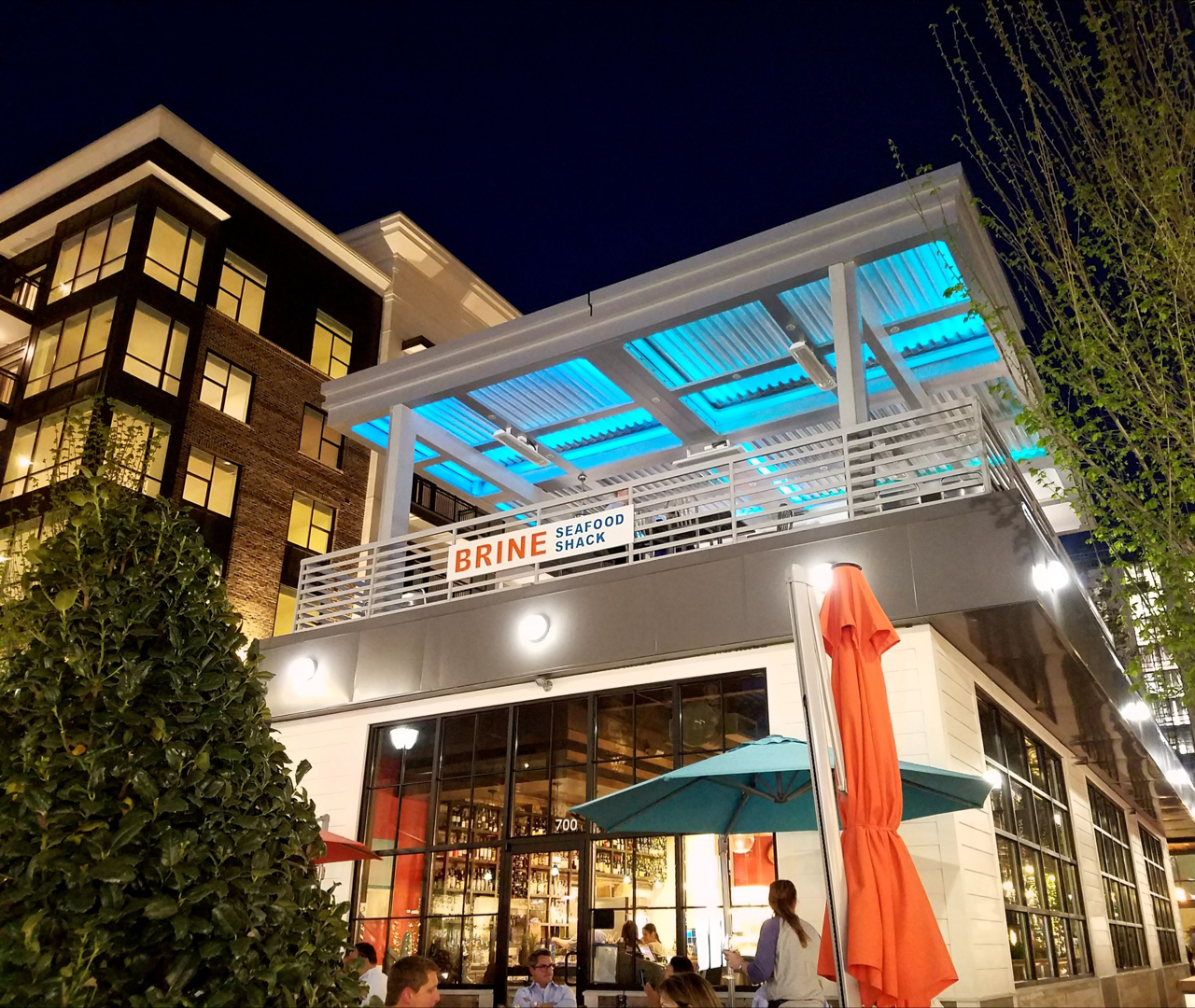 Night view of a seafood shack named 'Brine' with outdoor patio, blue lighting, and people dining outside.