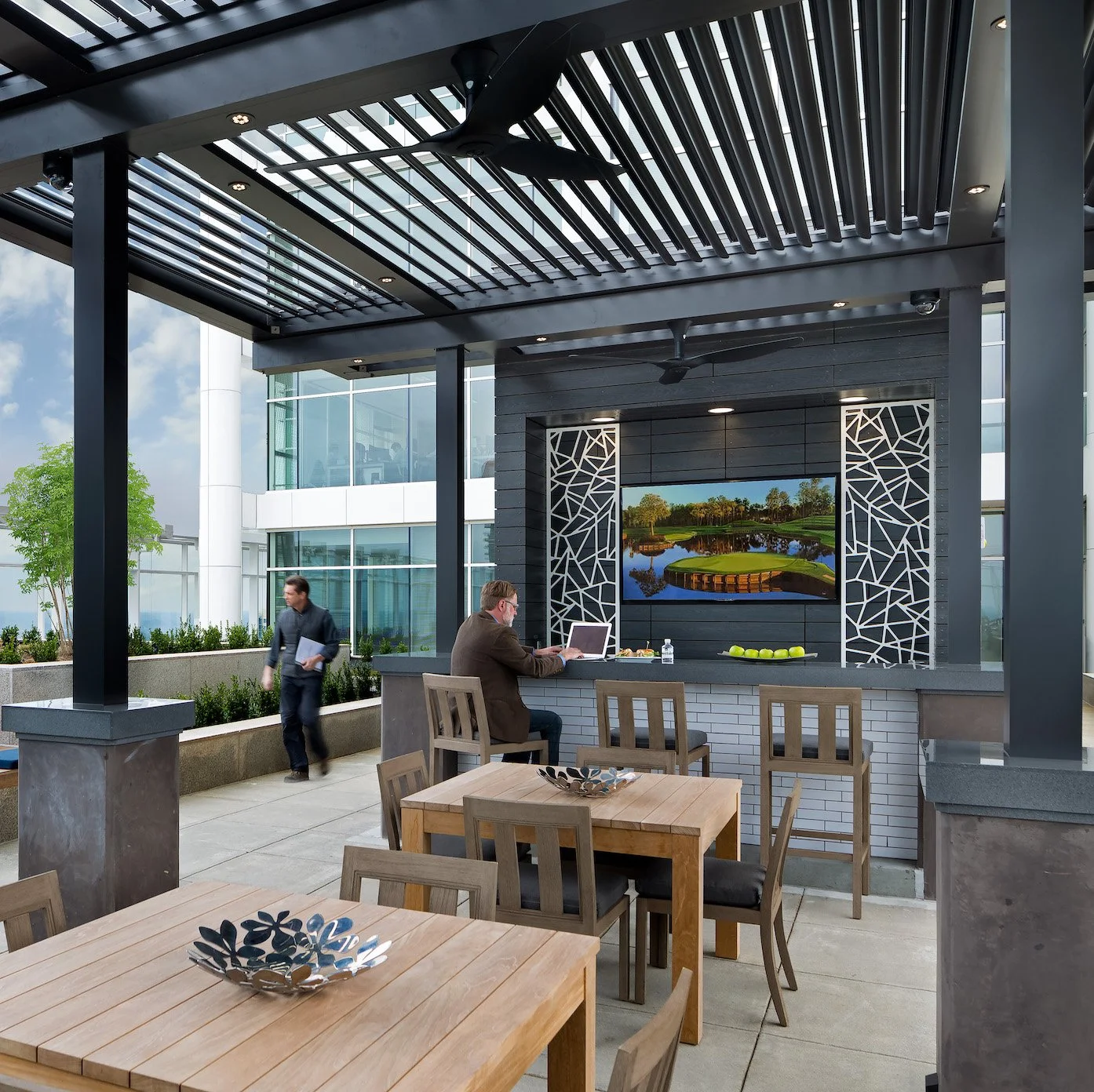 Outdoor rooftop lounge area with wooden tables and chairs, a large flat-screen TV showing a golf course, and two men working on a laptop and walking, with a modern building in the background.