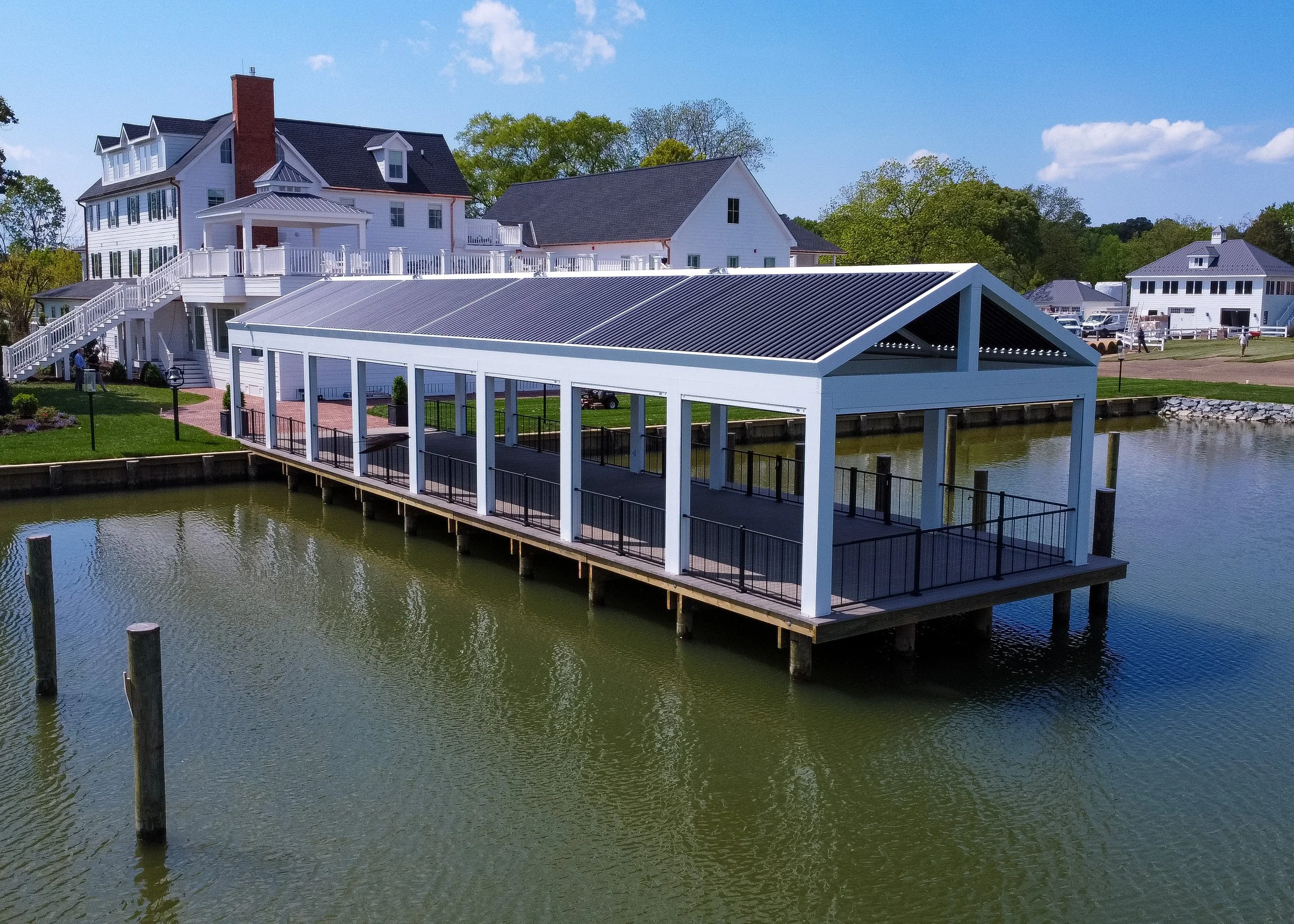 A waterfront scene featuring a white pavilion with a metal roof on a wooden dock over water, with houses and greenery in the background on a sunny day.