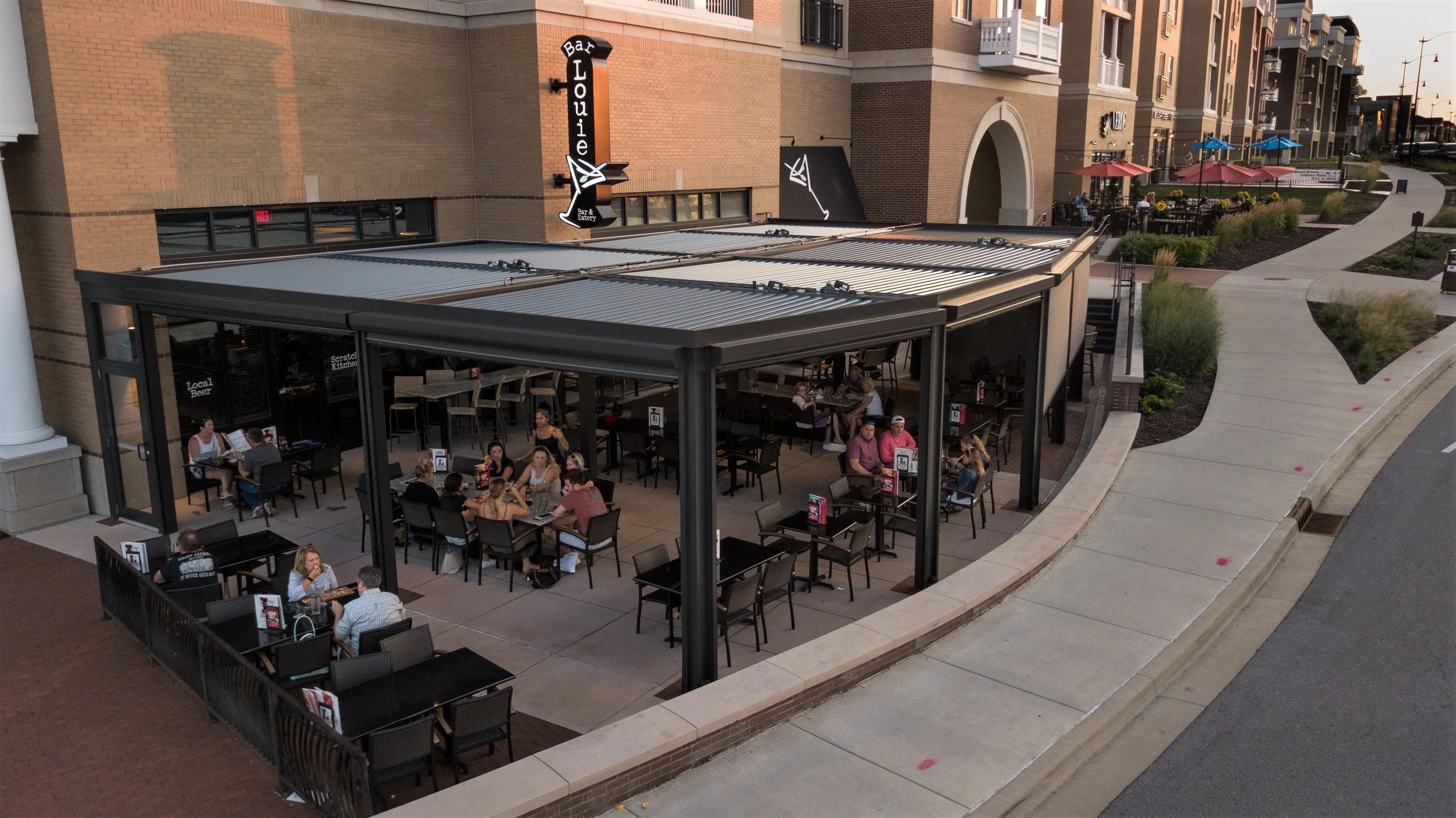 Outdoor patio dining area with multiple tables and people, adjacent to a bar called Louie's, situated next to a building with a curved sidewalk, some plants, and outdoor seating with umbrellas.