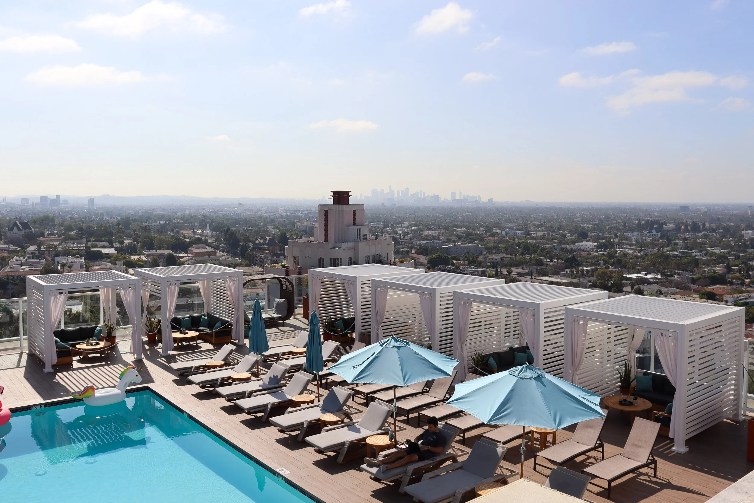Rooftop pool area with lounge chairs, umbrellas, cabanas with curtains, and a city skyline in the background on a sunny day.